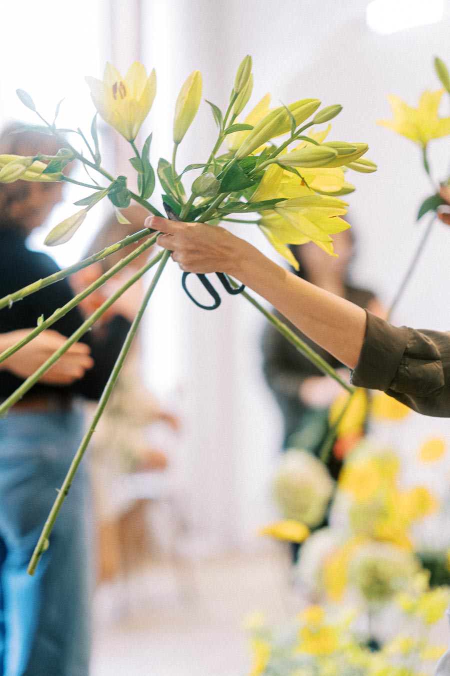 Two people exchanging a bouquet of vibrant yellow lilies in a bright, indoor setting, emphasizing a gesture of giving or receiving flowers, with blurred figures in the background enhancing a sense of depth and focus.