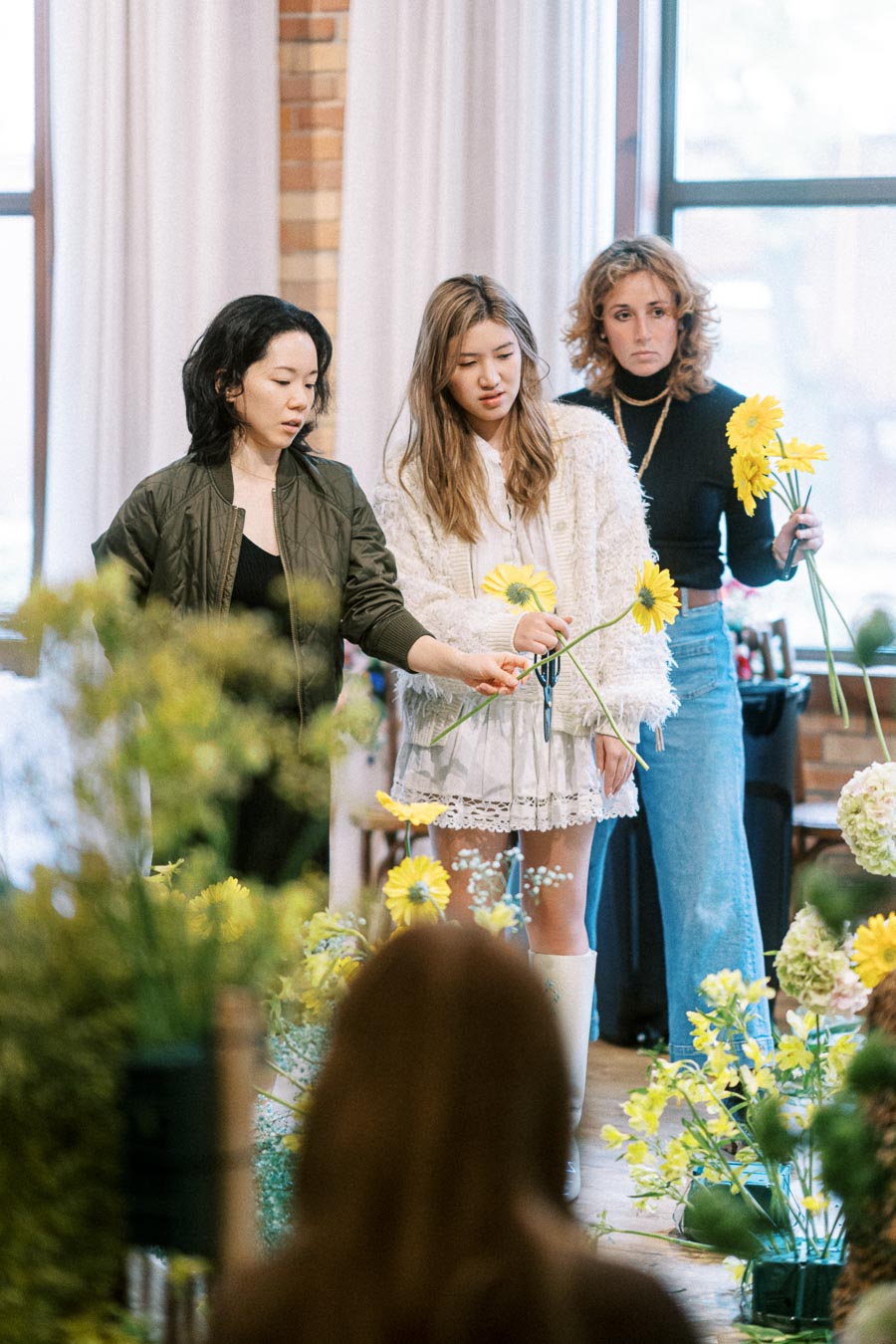 Three women engaging in flower arrangement, selecting yellow flowers in a sunlit room with brick walls and white curtains.