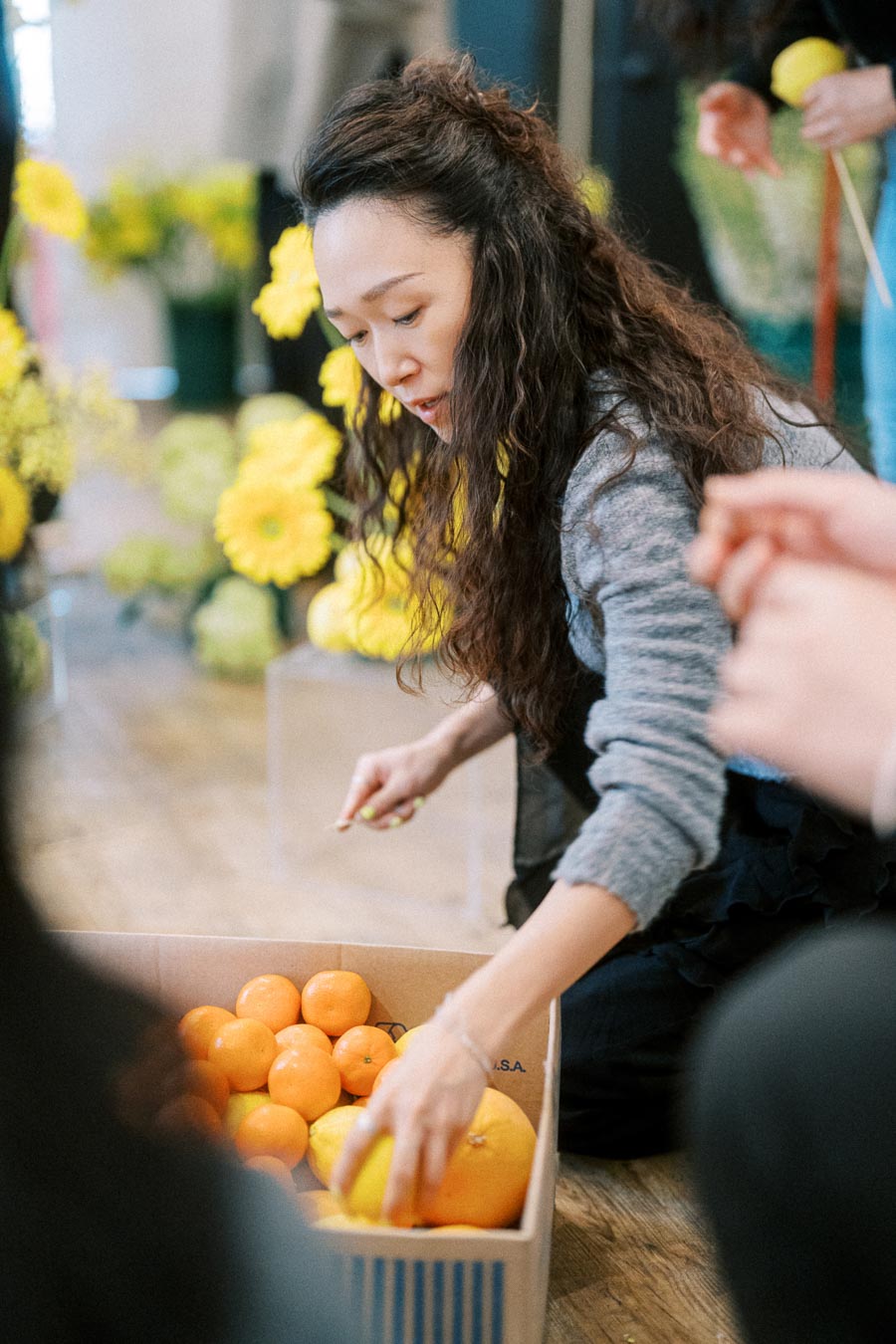 Woman arranging fresh citrus fruits in a box, surrounded by vibrant yellow flowers indoors.