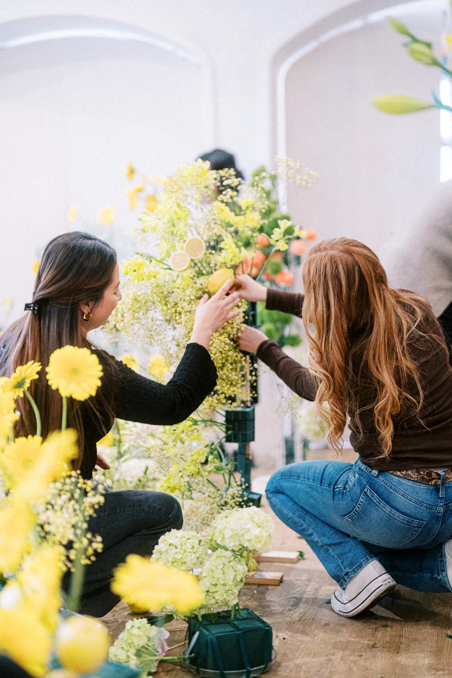 Two women crafting a decorative floral arrangement with yellow and green flowers and citrus accents, inside a bright room, showcasing creativity and teamwork.