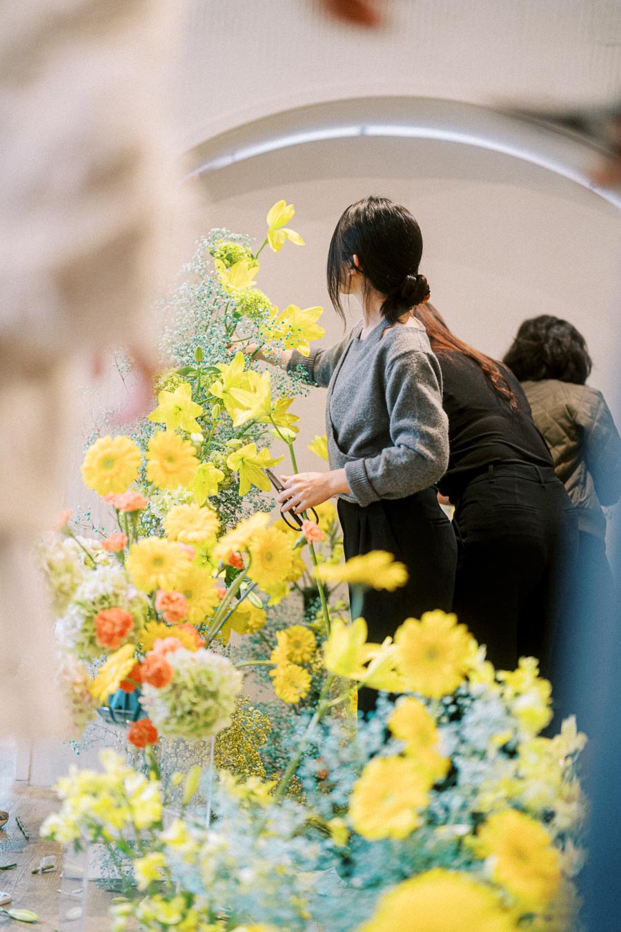 A woman arranging vibrant yellow flowers in a floral display inside a bright room, emphasizing botanical elegance and creative gardening.