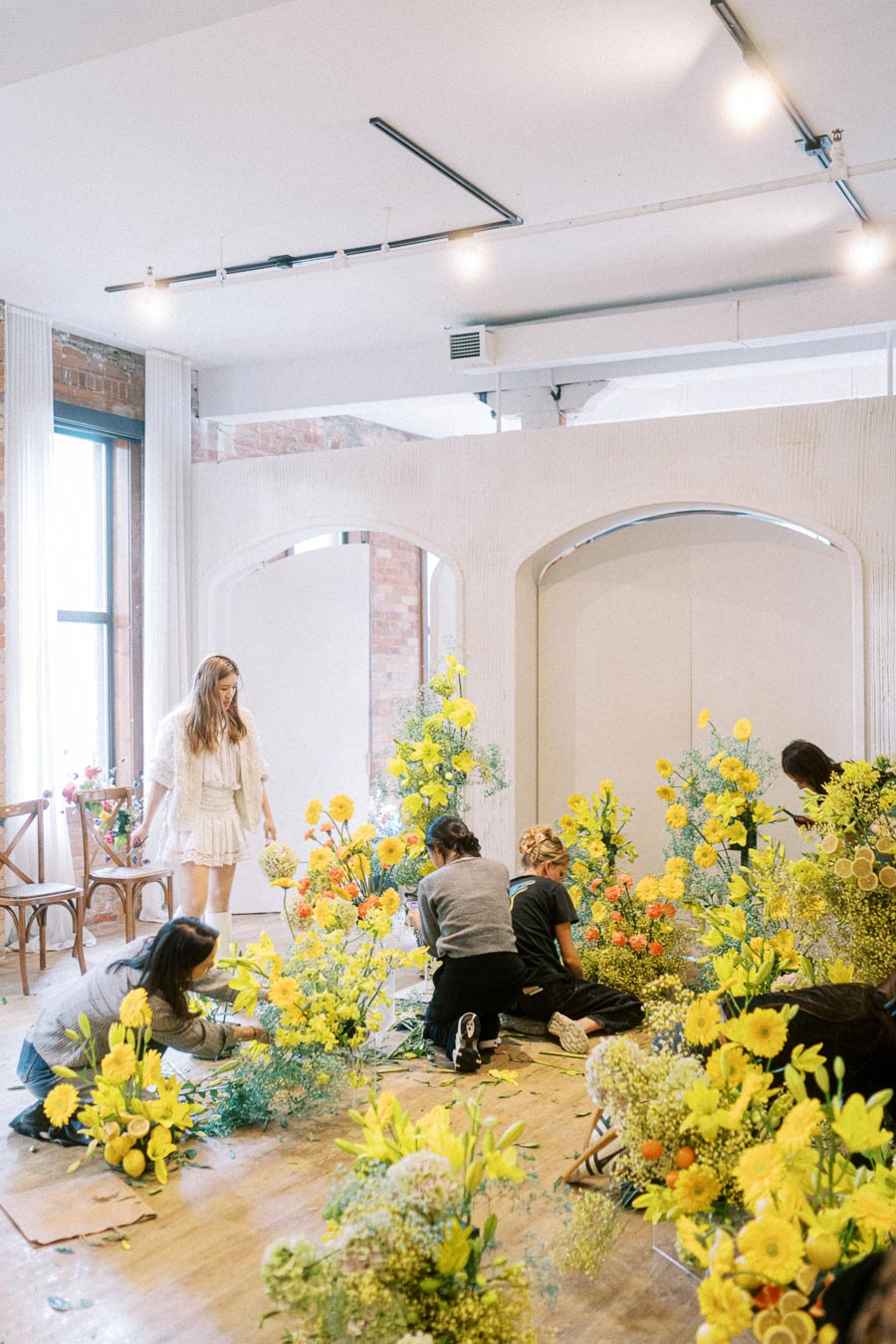 Group of people arranging vibrant yellow and orange flowers in a bright, modern interior space, with natural light streaming through large windows.