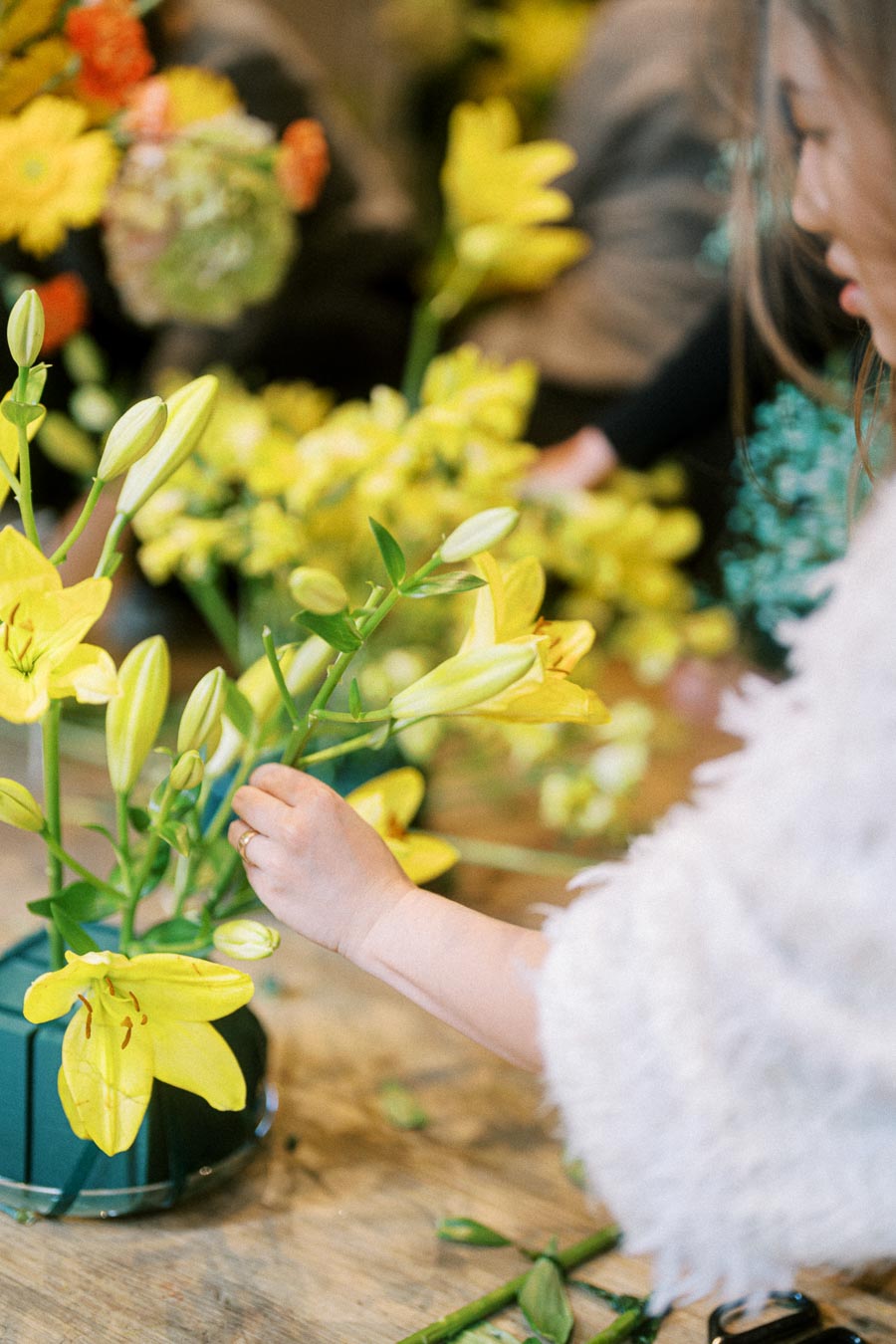 Child arranging vibrant yellow lilies in a floral workshop, surrounded by a variety of colorful flowers on a wooden table.