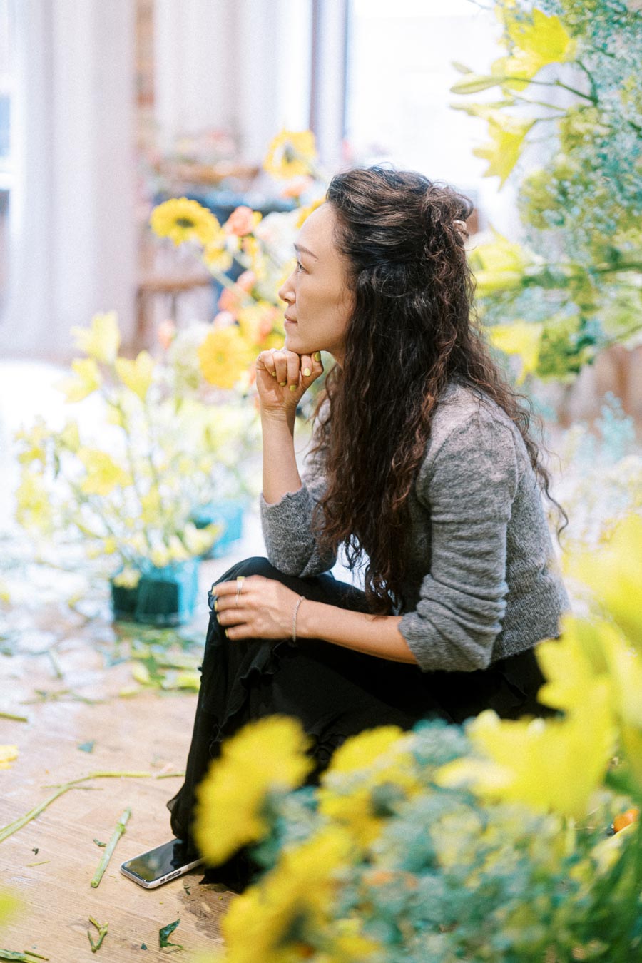 A woman with long, curly hair sitting thoughtfully among yellow flowers in a bright, cluttered room, wearing a grey sweater and black skirt; a smartphone rests on the floor nearby.