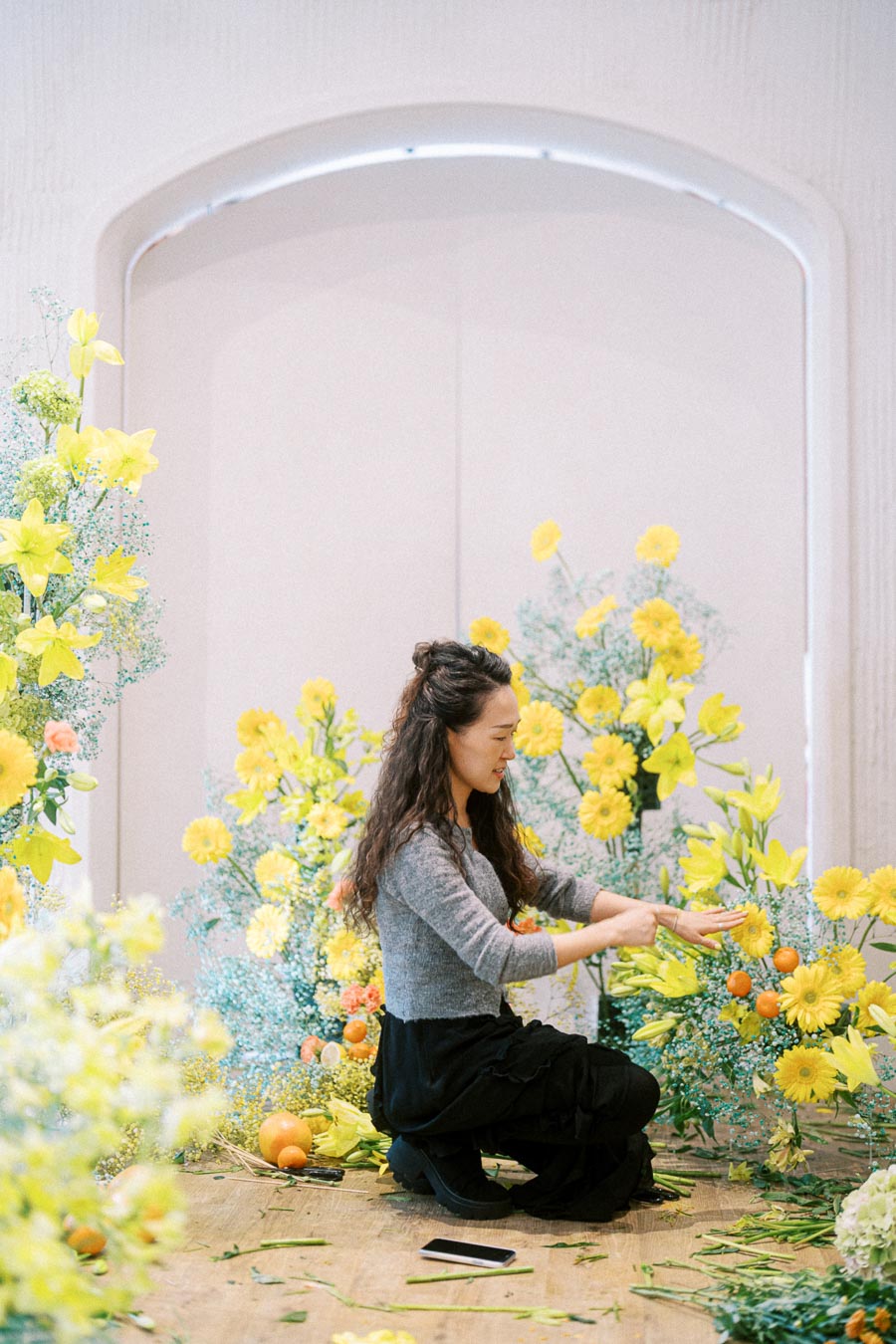 Florist arranging a vibrant display of yellow flowers and oranges in a bright room, demonstrating floral arrangement techniques.