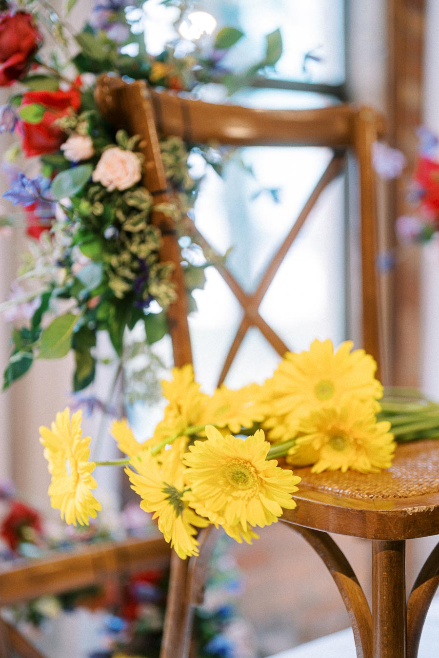 A wooden chair adorned with vibrant yellow gerbera daisies and an arrangement of colorful flowers, creating an elegant and festive atmosphere in a bright, sunlit setting.