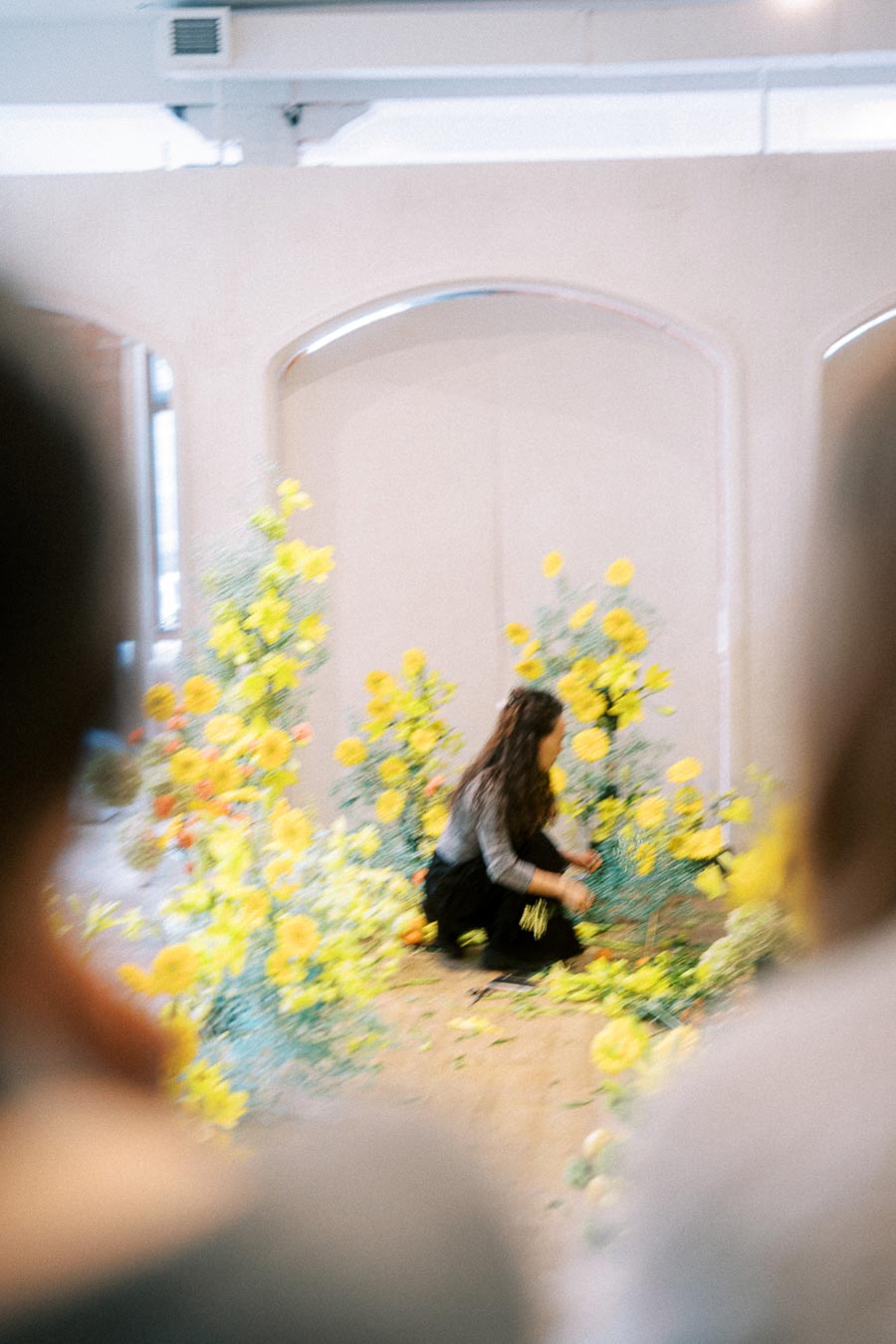 Blurry image of a woman arranging vibrant yellow and orange flowers in an indoor setting, creating a colorful floral display amidst soft lighting.