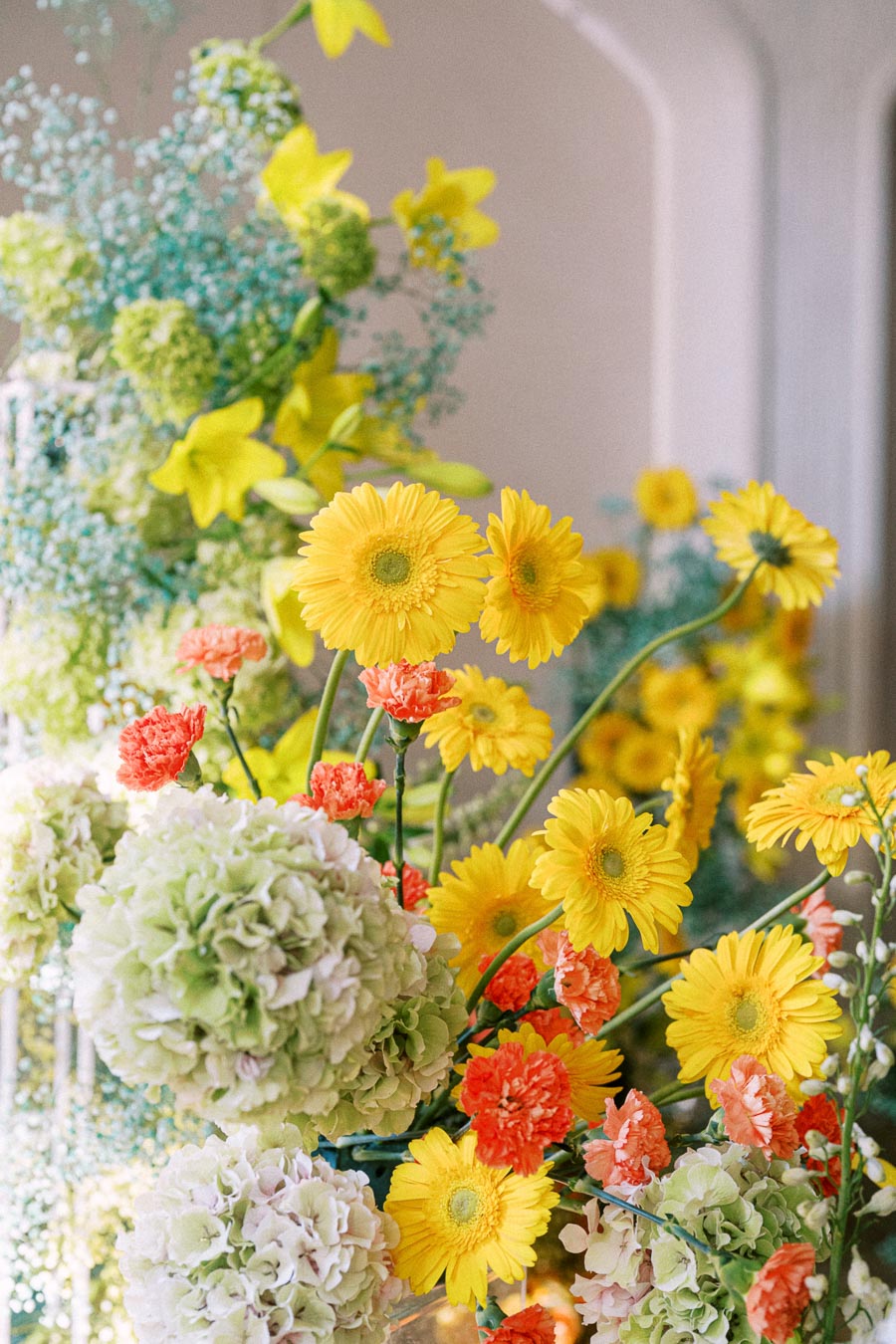 A vibrant floral arrangement featuring yellow gerbera daisies, orange carnations, and green hydrangeas set against a soft-focus background. Ideal for spring wedding decor or garden-themed events.