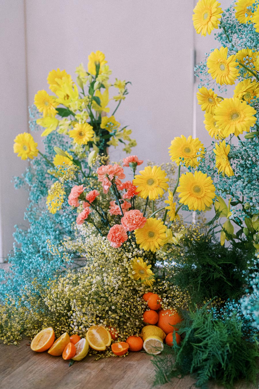 Vibrant floral arrangement with yellow daisies, pink carnations, and lush green foliage, surrounded by fresh oranges, lemons, and sliced citrus fruits on a wooden floor.