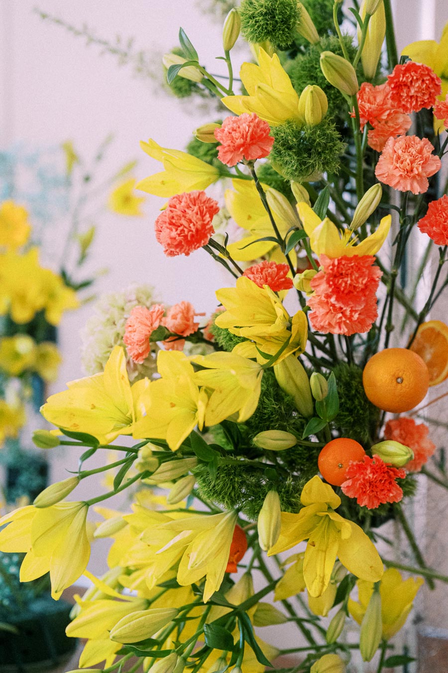 Bright and colorful floral arrangement with yellow lilies, orange carnations, and fresh oranges, creating a vibrant and lively display.