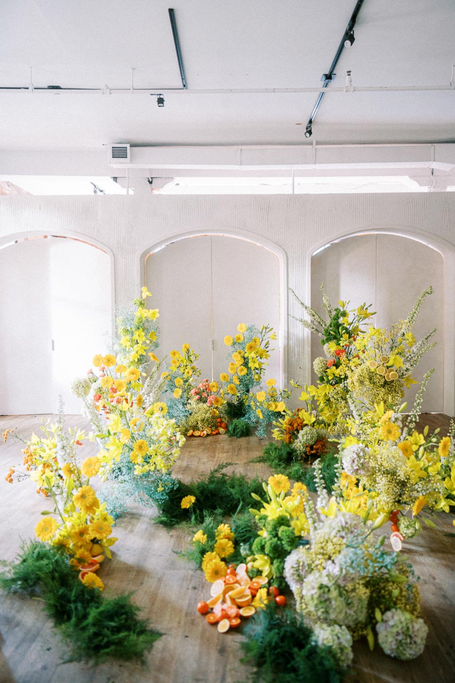 Vibrant floral installation with yellow flowers and greenery, surrounded by assorted citrus fruits on a wooden floor, set against a minimalist white backdrop.
