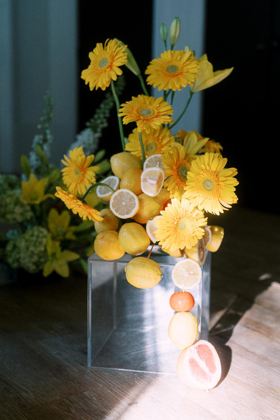 A creative floral arrangement featuring vibrant yellow daisies and sliced lemons in a clear vase, artistically displayed in natural sunlight on a wooden table.