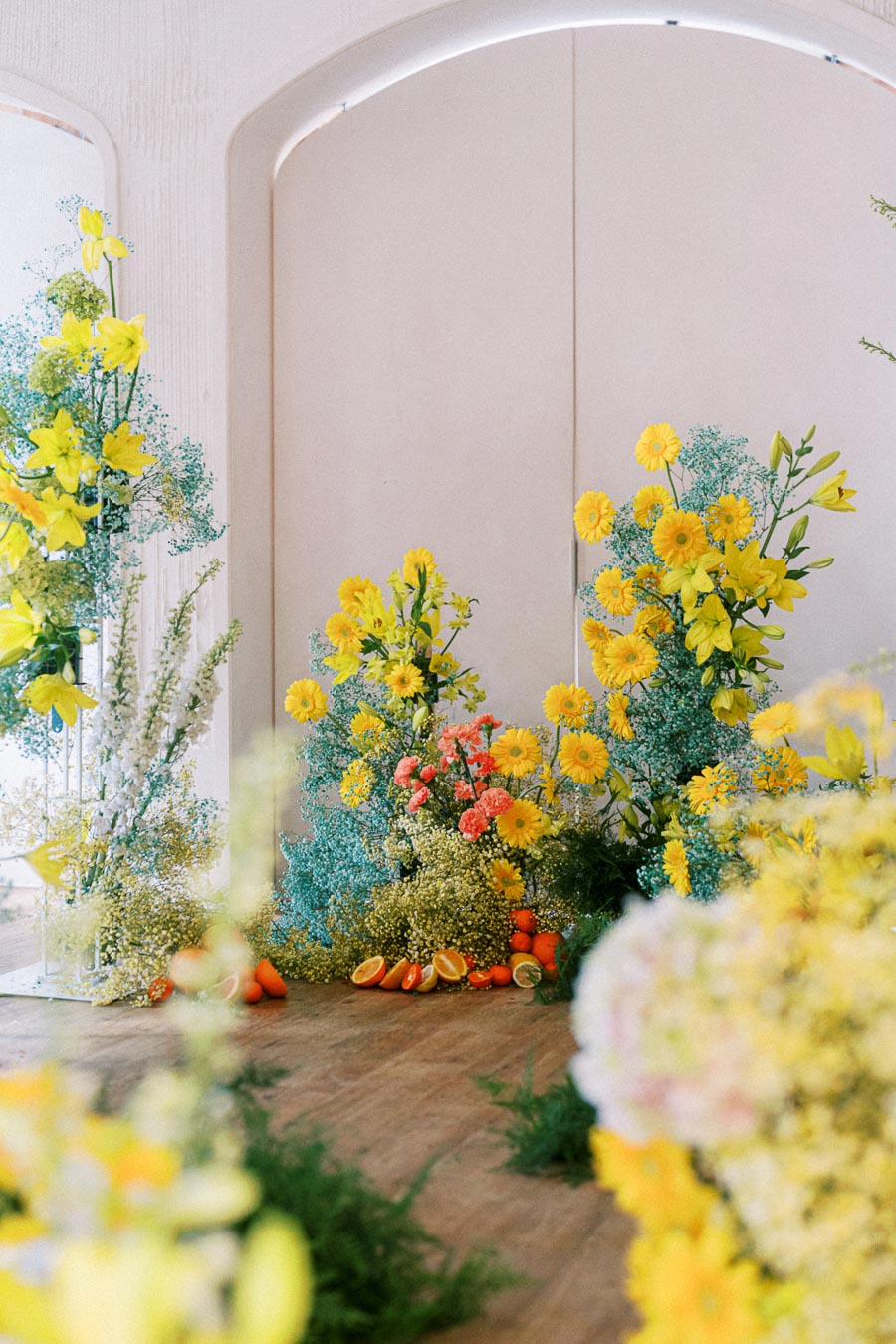 Vibrant floral arrangement with yellow and orange flowers, blue accents, and citrus fruits, set against a light-colored archway backdrop.
