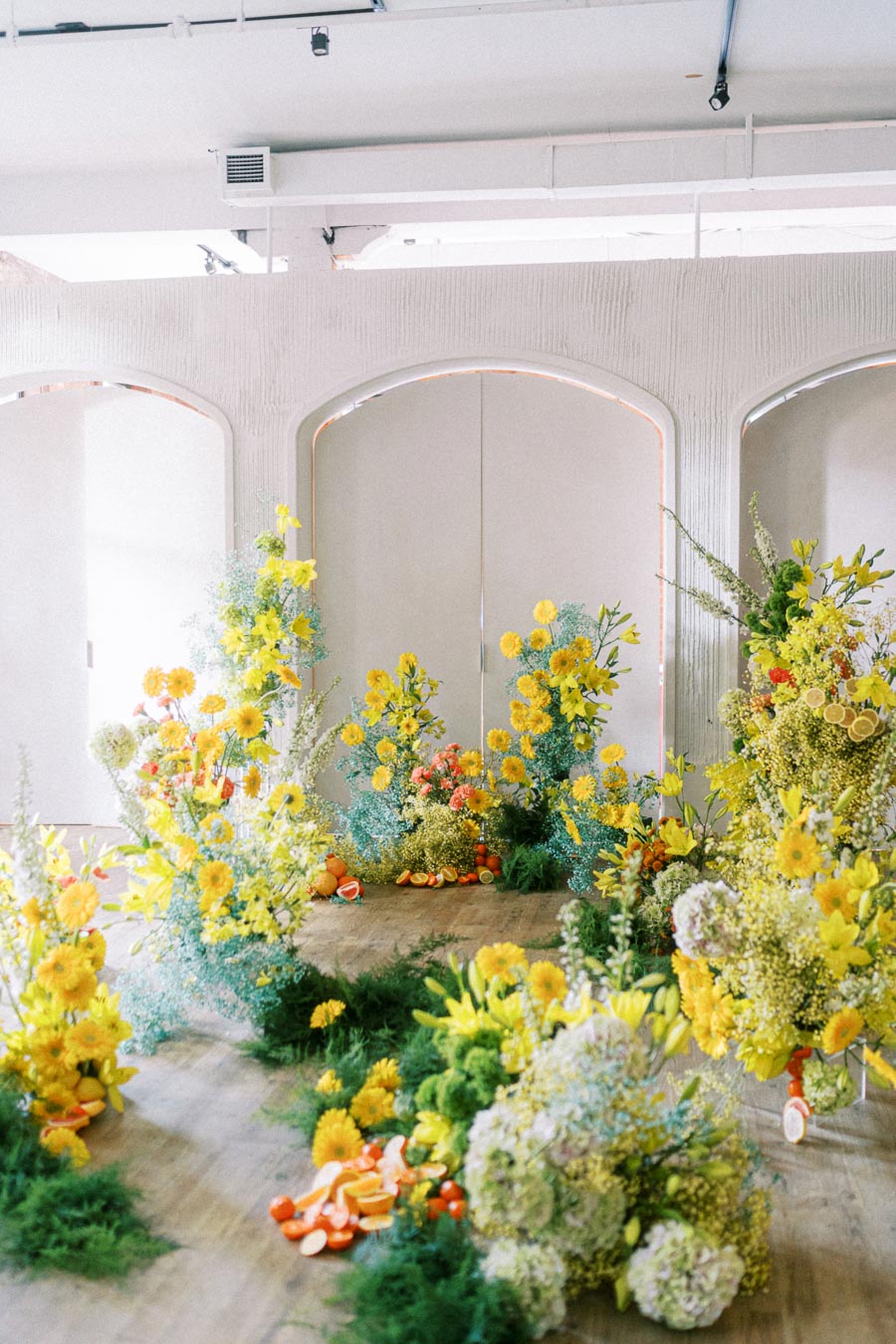 Vibrant floral arrangement featuring yellow and green flowers with a scattering of orange slices on a wooden floor, set in a light-filled modern venue.