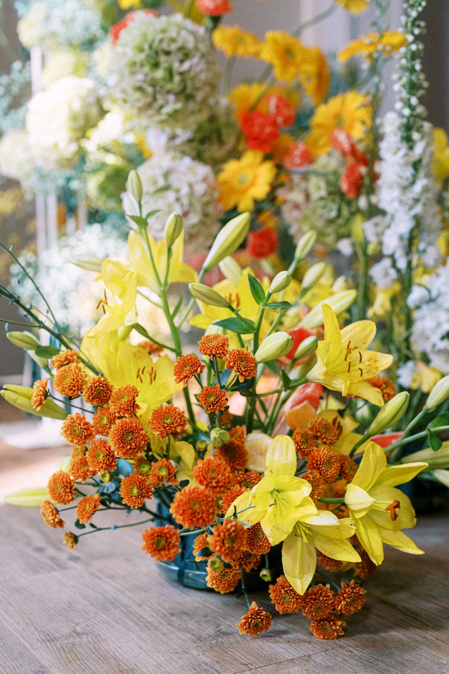 Vibrant floral arrangement with yellow lilies and orange chrysanthemums on a wooden table, surrounded by diverse colorful blooms in the background.