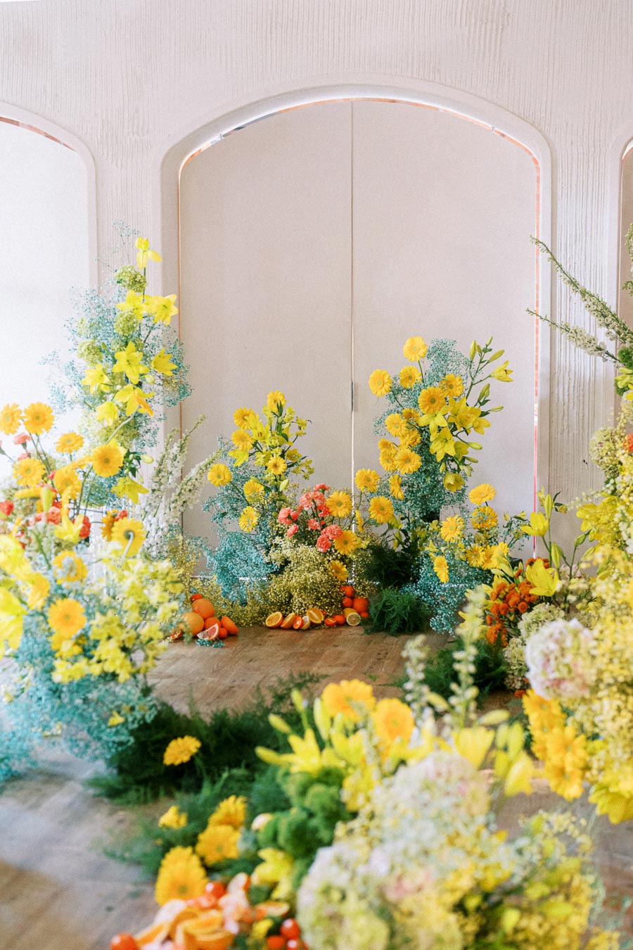 Vibrant floral arrangement featuring yellow daisies and lilies, adorned with greenery and accompanied by fresh oranges and citrus slices, set against a textured white wall.