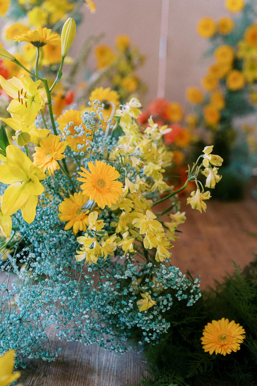 Bright and colorful floral arrangement featuring yellow daisies and lilies mixed with delicate blue baby's breath, set against a soft-focus background of similar flowers.
