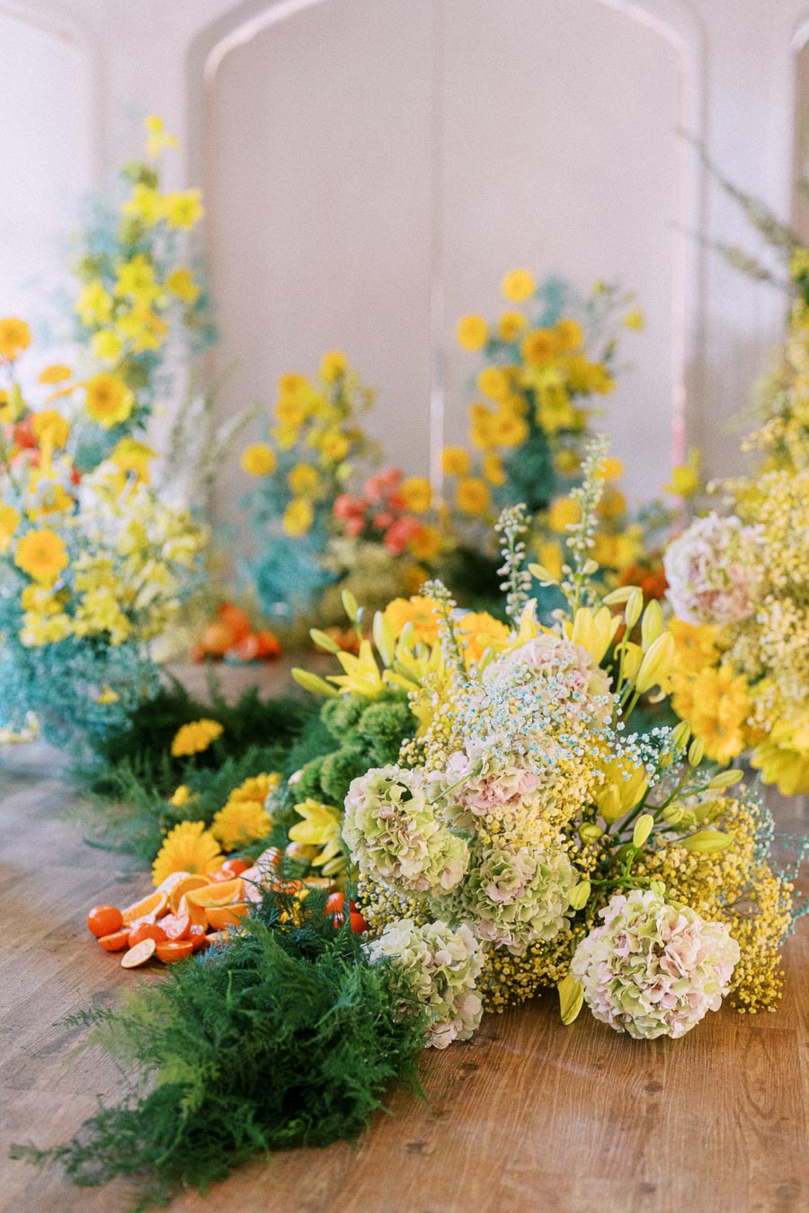 Colorful floral arrangement with yellow daisies, green foliage, and pastel hydrangeas on a wooden floor, accented by orange fruits, creating a vibrant and fresh decor.