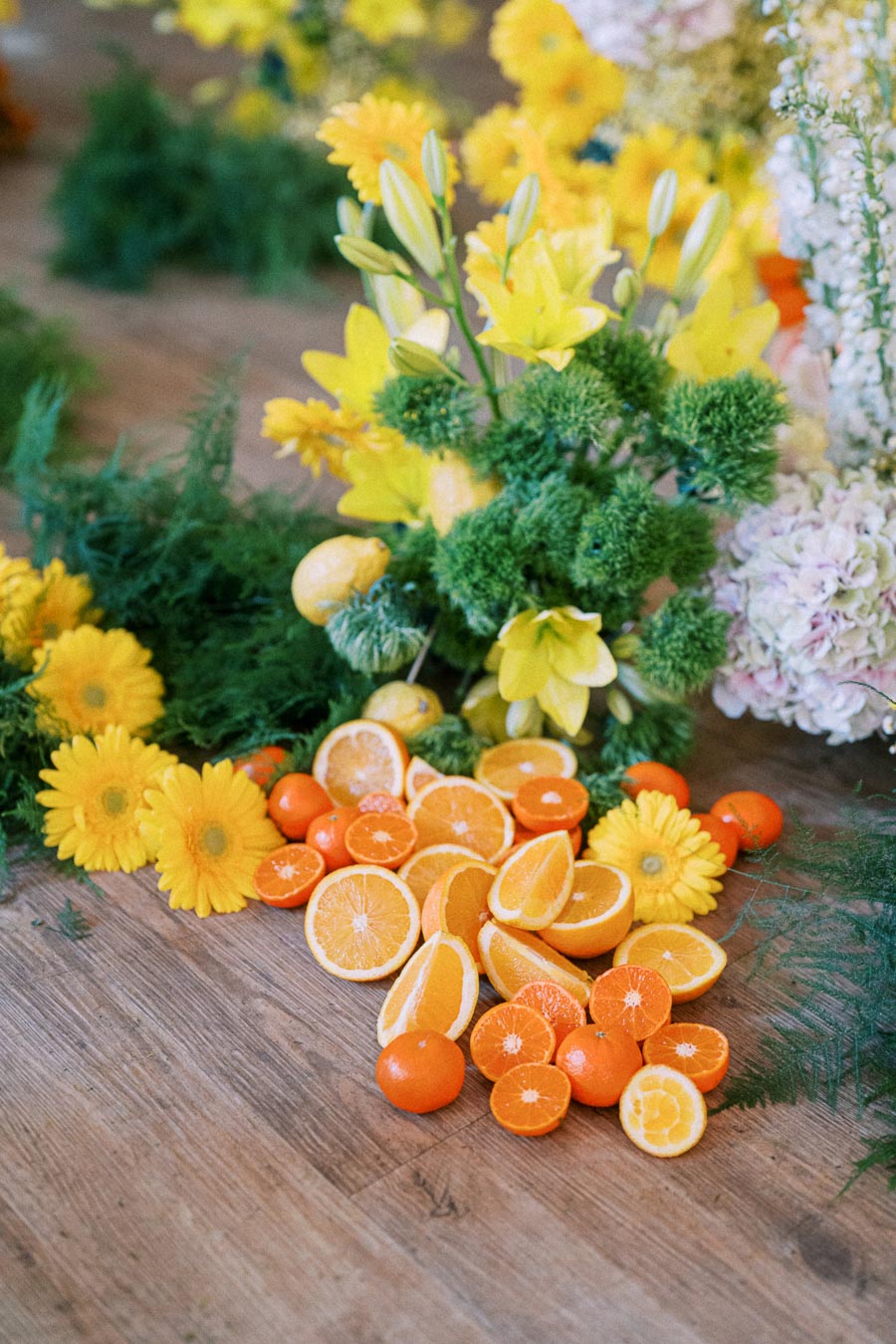 A vibrant arrangement of fresh sliced oranges and whole tangerines surrounded by bright yellow flowers and lush greenery on a wooden surface, showcasing a cheerful and natural aesthetic.