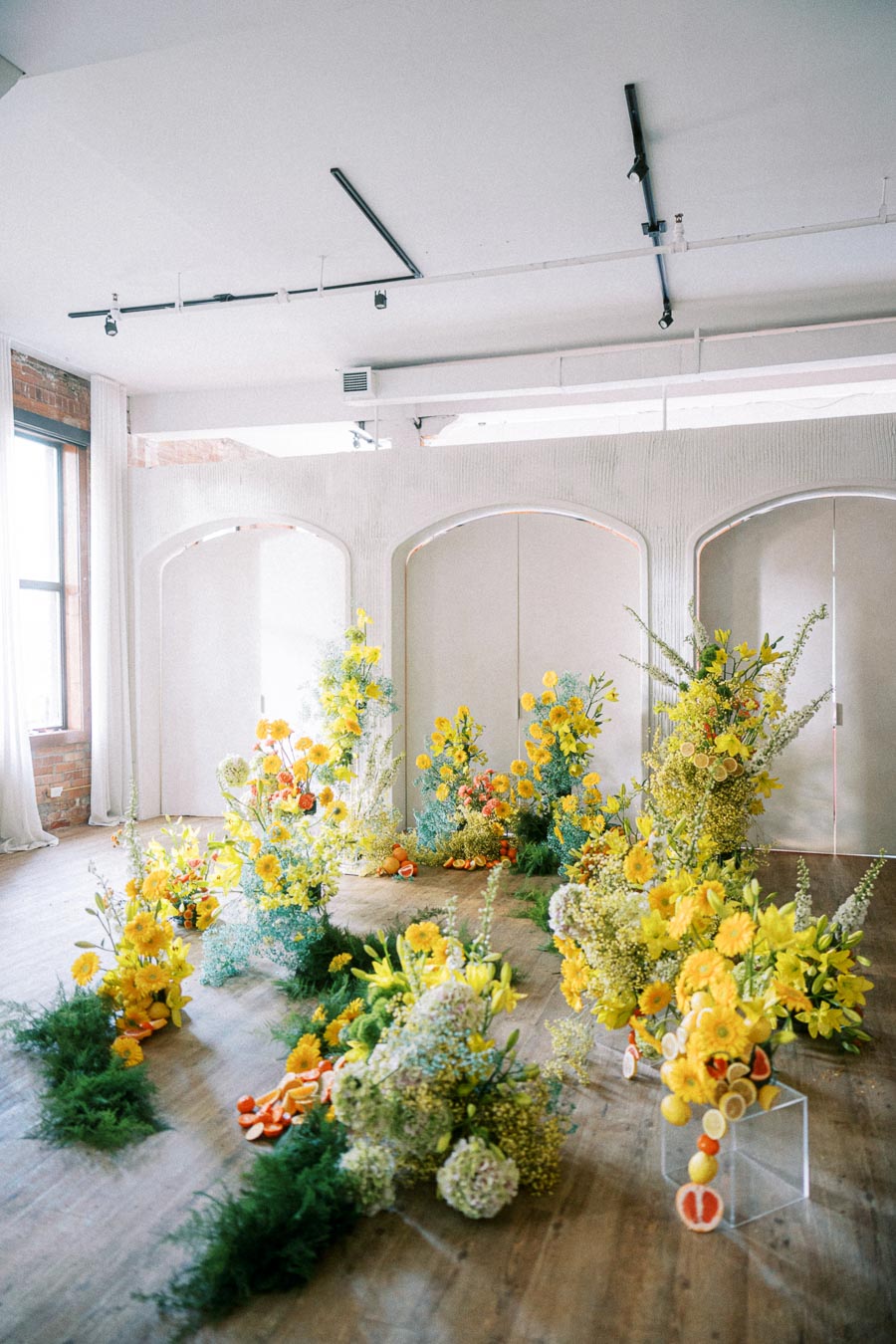 A bright and airy indoor arrangement showcasing vibrant yellow and orange floral displays mixed with lush greenery, set against a modern studio backdrop with large windows and wooden flooring.