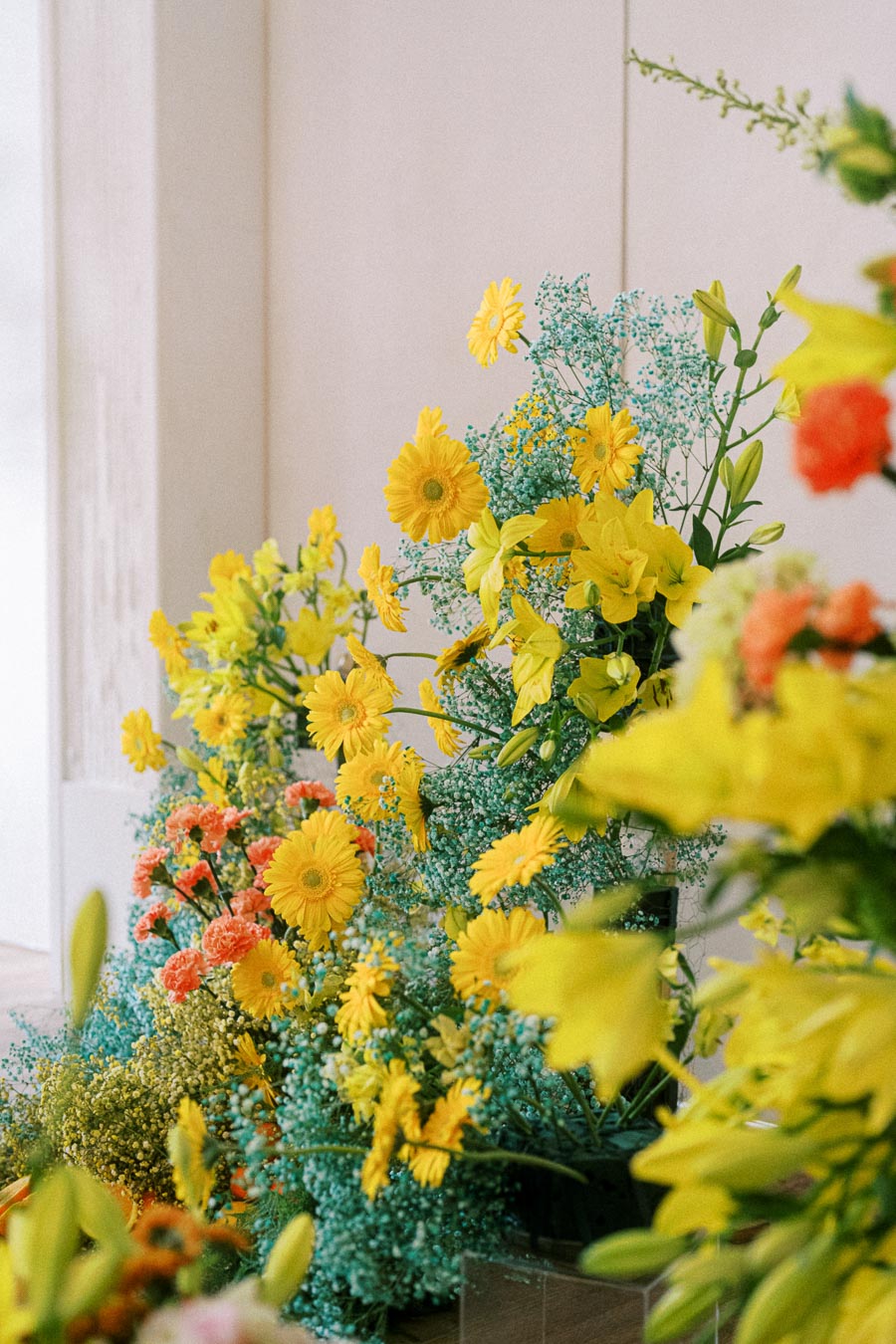 Floral arrangement featuring vibrant yellow daisies, lilies, and bright orange carnations, surrounded by delicate blue baby's breath, set against a soft neutral background.
