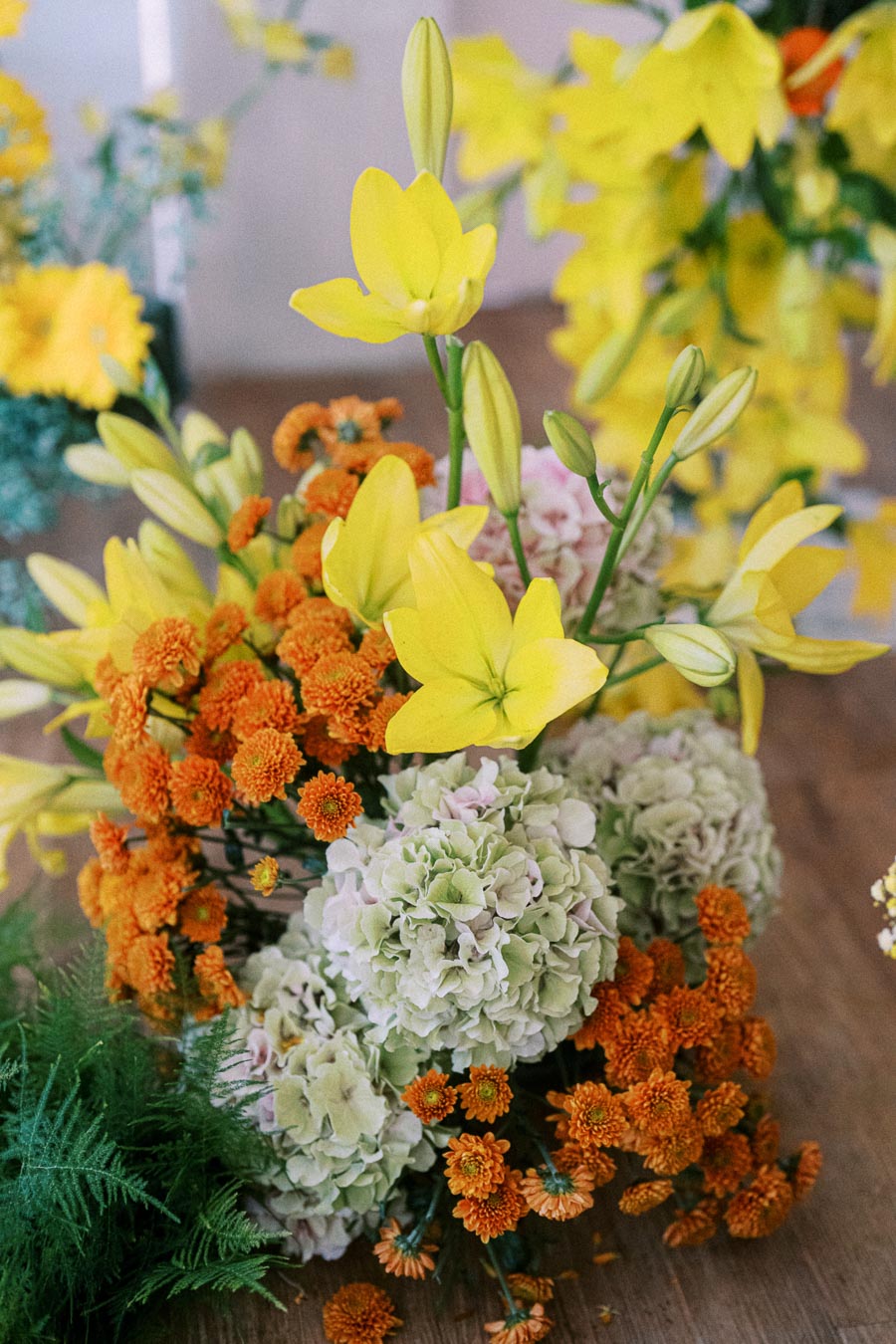 A vibrant floral arrangement featuring bright yellow lilies, soft green hydrangeas, and clusters of small orange chrysanthemums, displayed on a wooden table with lush green foliage in the background. Perfect for spring or summer decor inspiration.