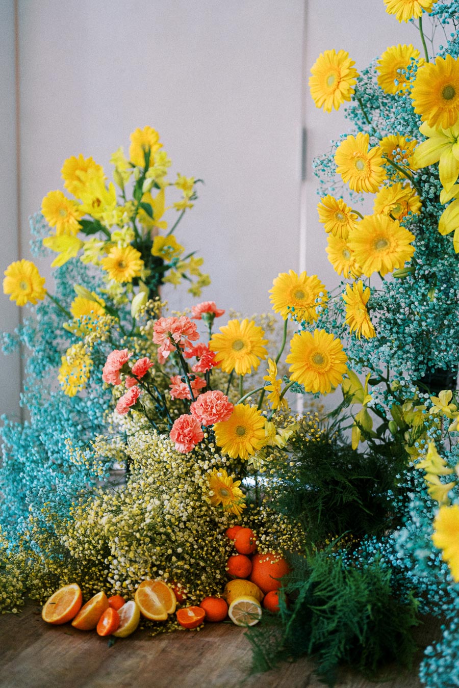 Vibrant floral arrangement with yellow daisies, pink carnations, and blue baby's breath, accented by fresh citrus fruits including oranges and lemons, on a wooden surface.