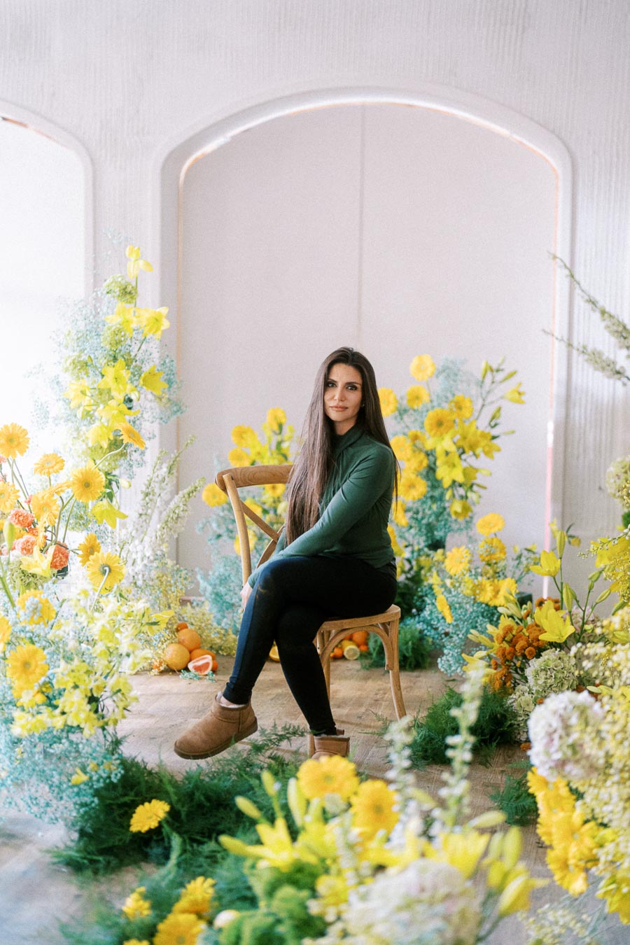 A woman sitting on a wooden chair surrounded by vibrant yellow flowers and greenery, creating a serene and colorful indoor garden setting.