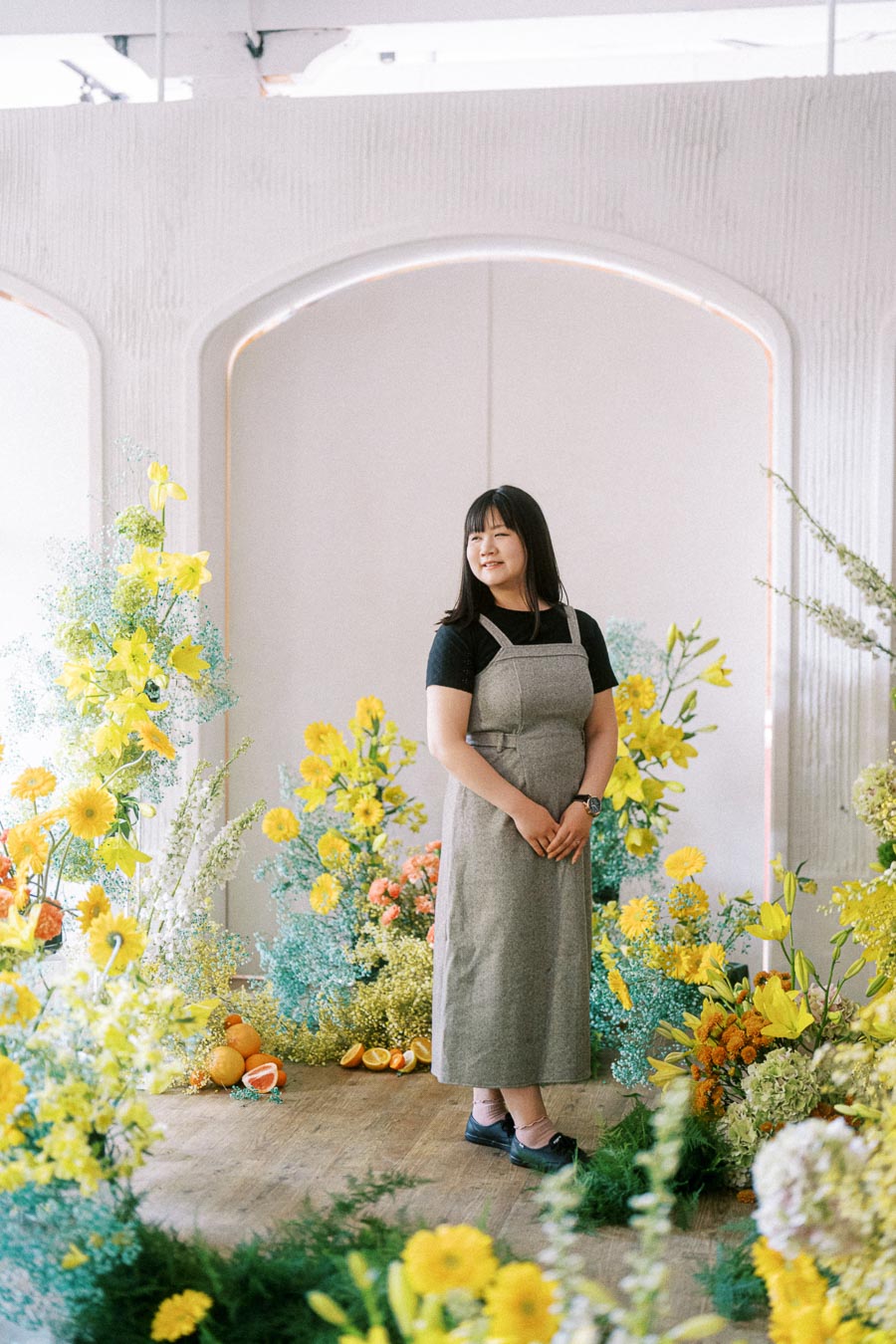 Young woman in a gray dress standing in a vibrant floral arrangement with yellow and orange flowers, surrounded by a modern, curved architectural backdrop.