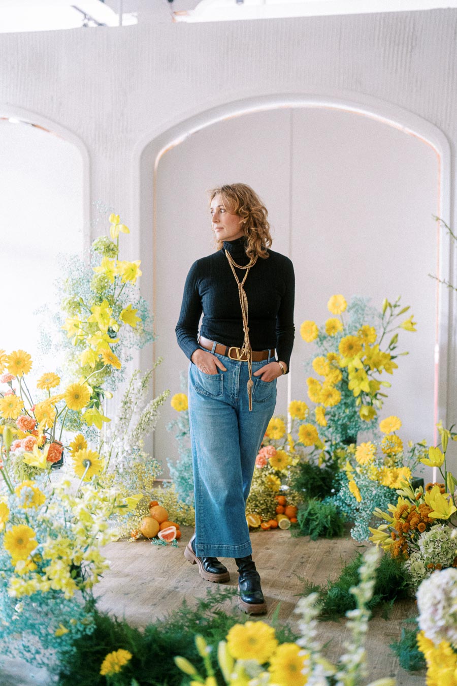 Woman in a black turtleneck and jeans stands amidst vibrant yellow flowers and greenery in a brightly lit room.