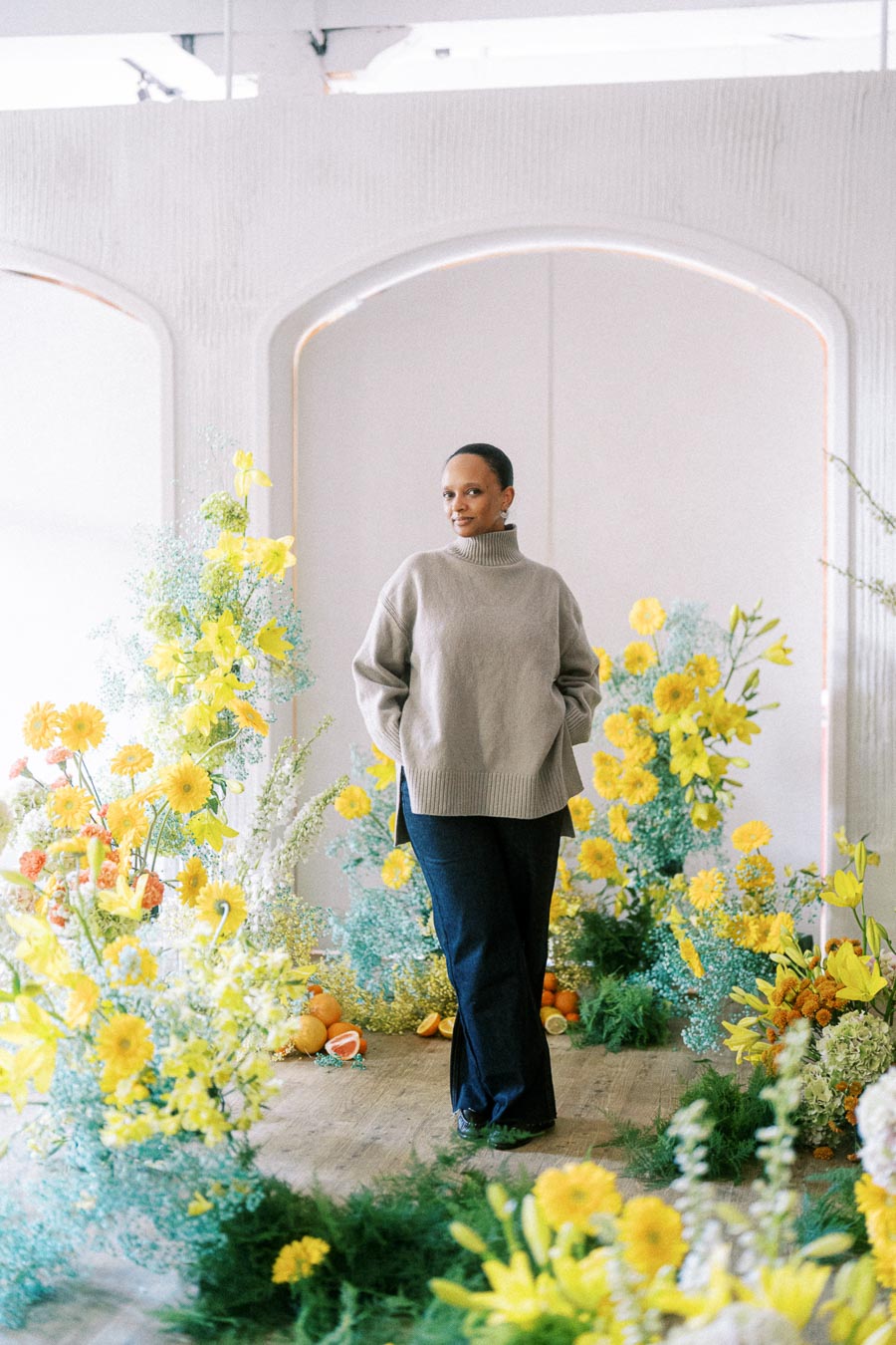 A person wearing a cozy beige sweater and dark jeans stands confidently among vibrant yellow flowers and green foliage inside a bright, modern room with arched windows.