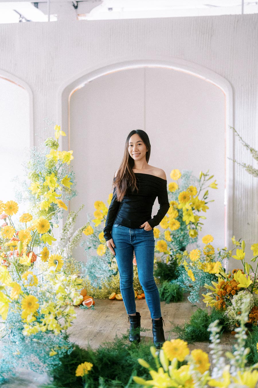 A woman in a black off-the-shoulder top and jeans stands amidst a vibrant floral display featuring yellow and orange flowers and greenery inside a bright room.
