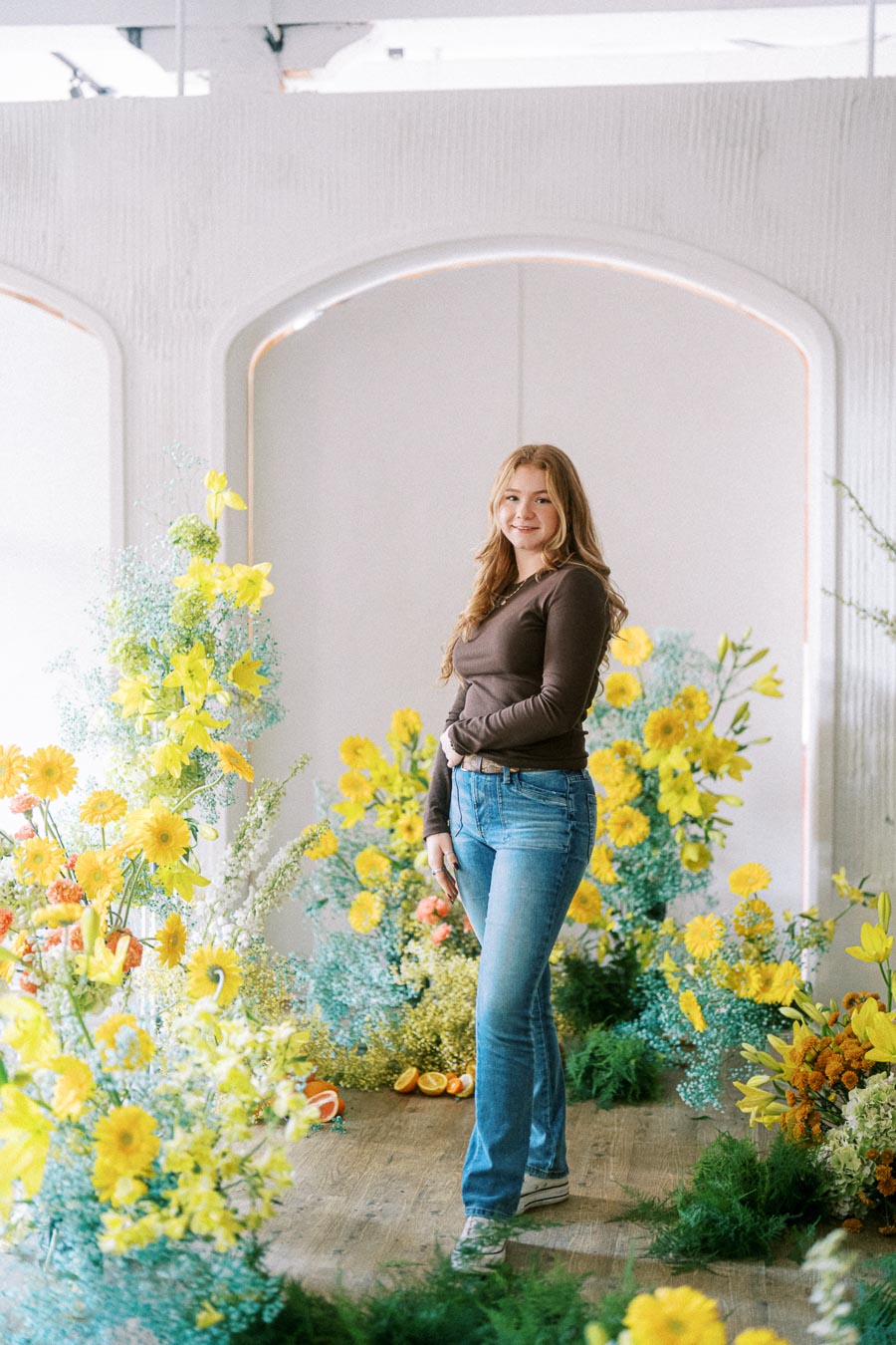 A woman in casual attire stands in a bright room surrounded by vibrant yellow flowers and greenery, creating a fresh and cheerful atmosphere.