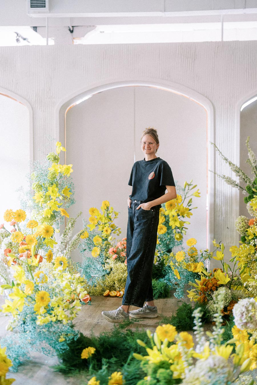 A person in casual attire stands amidst a vibrant floral arrangement featuring yellow and orange flowers, set against an elegant, arching backdrop inside a bright, modern space.