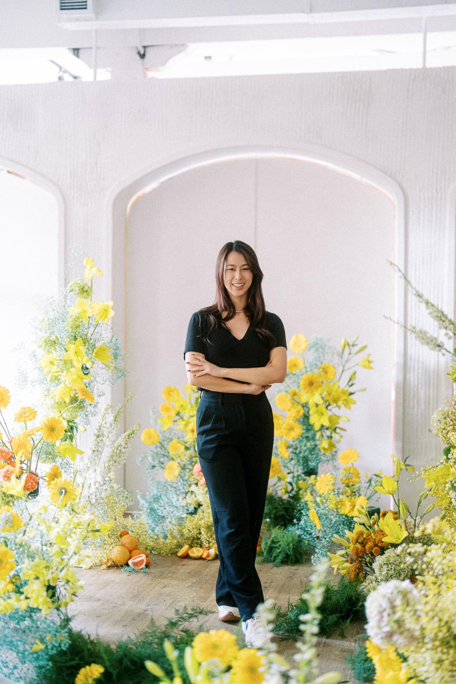 Woman in a black outfit standing amidst a vibrant floral setting with yellow and orange flowers, in a bright indoor space with natural light.