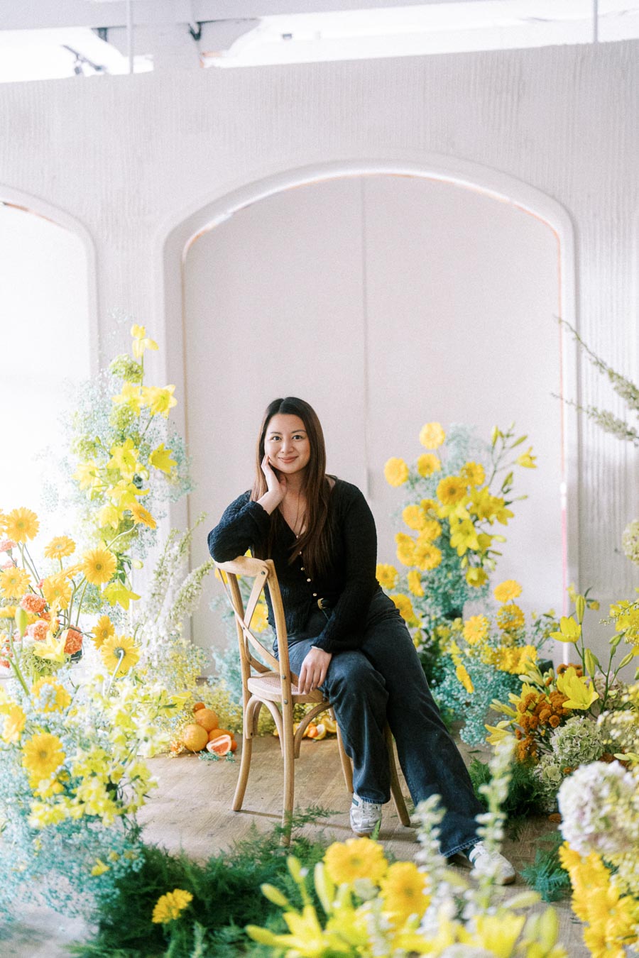 A woman sits on a wooden chair surrounded by vibrant yellow and orange flowers in a bright, airy room.