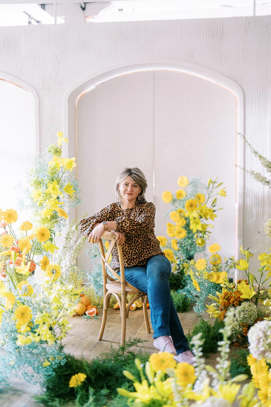 A person sitting on a wooden chair surrounded by vibrant yellow and orange flowers, wearing a leopard print blouse and jeans, in a bright, artistic setting.