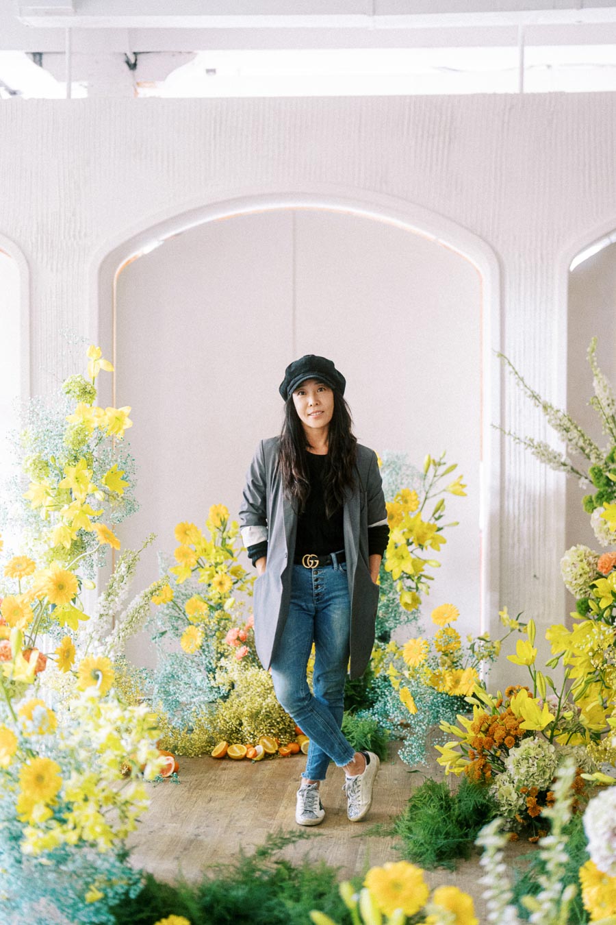 Woman in casual attire standing amidst a vibrant floral arrangement with yellow flowers, modern white archway in the background.