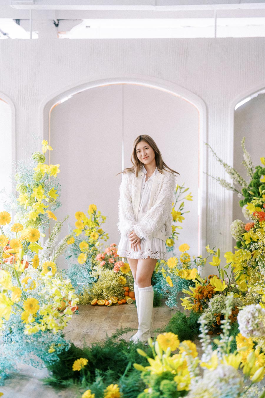A young woman in a white dress and boots stands surrounded by colorful flowers and greenery, creating a bright, cheerful atmosphere indoors.