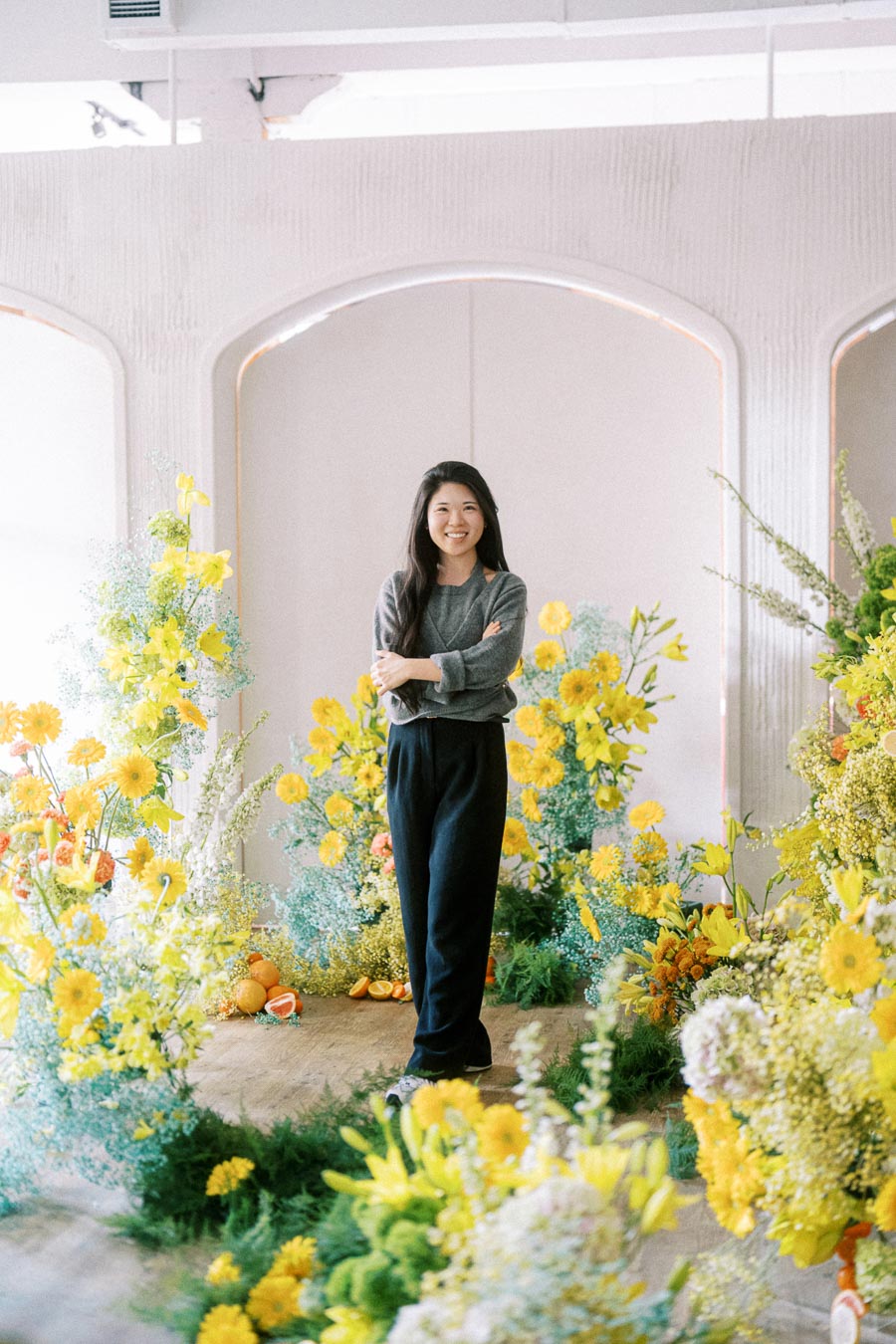 A person standing in a light-filled room surrounded by vibrant yellow and green floral arrangements, wearing a gray sweater and black pants, smiling at the camera.