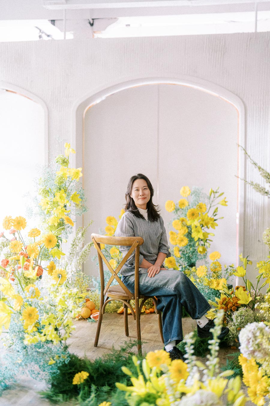 Woman seated on a wooden chair surrounded by vibrant yellow flowers and greenery in a bright, serene setting.