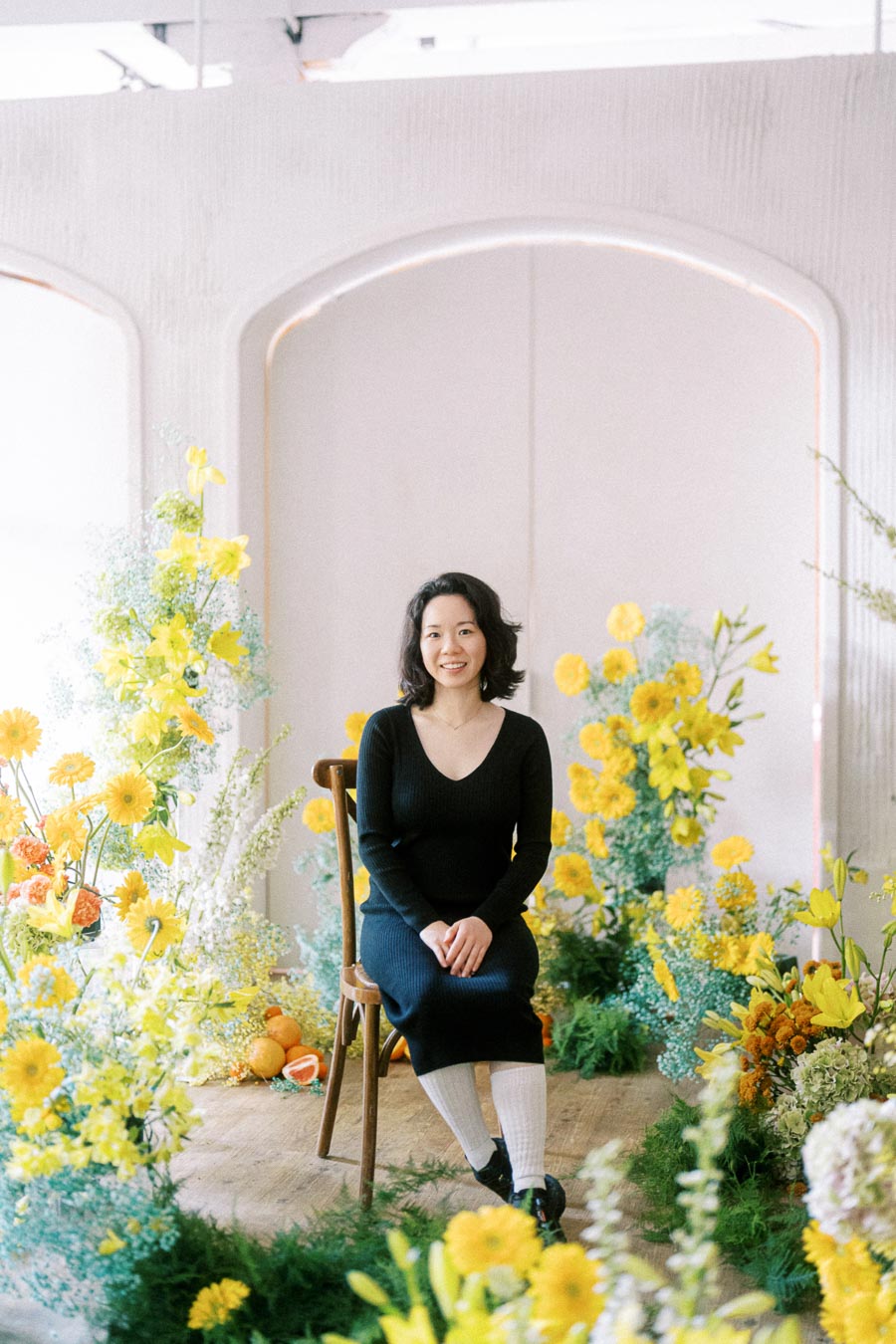 A person sitting on a wooden chair surrounded by vibrant yellow flowers and greenery, in a bright, elegantly decorated room.