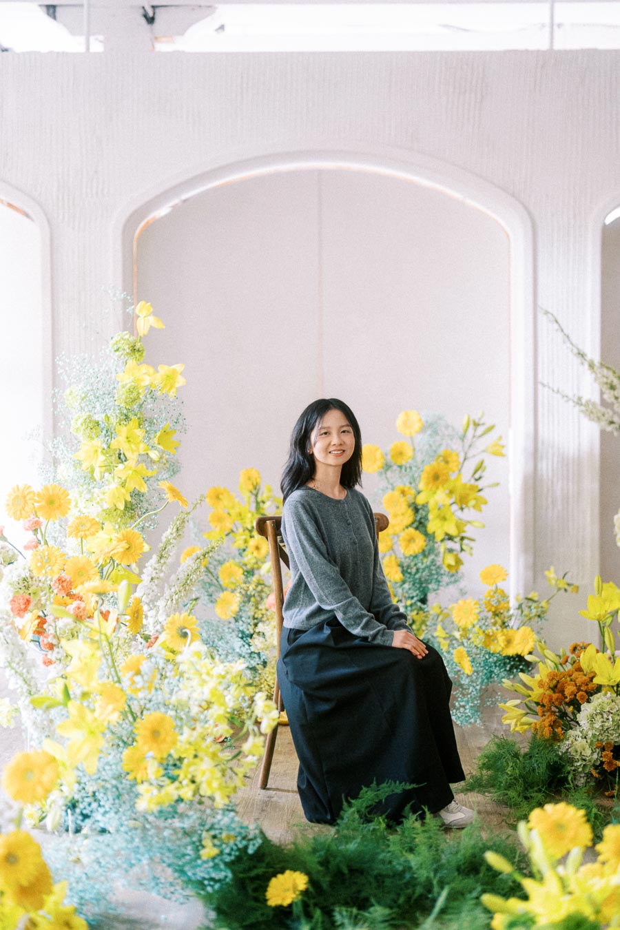 Young woman sitting on a chair surrounded by vibrant yellow flowers in a bright, minimalist setting.