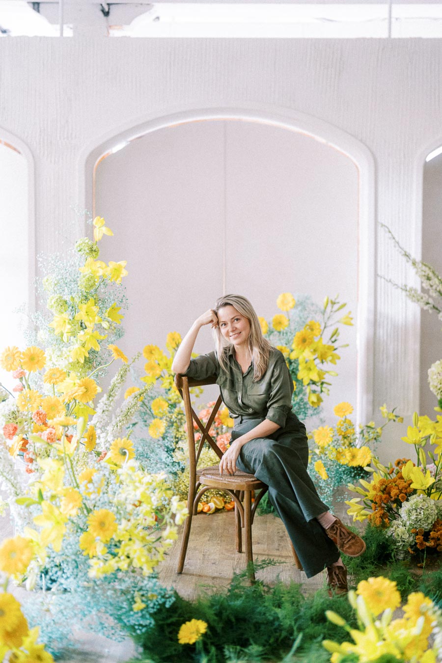 Woman seated amidst vibrant yellow and green floral display, smiling while wearing a green outfit, in a light-filled room.