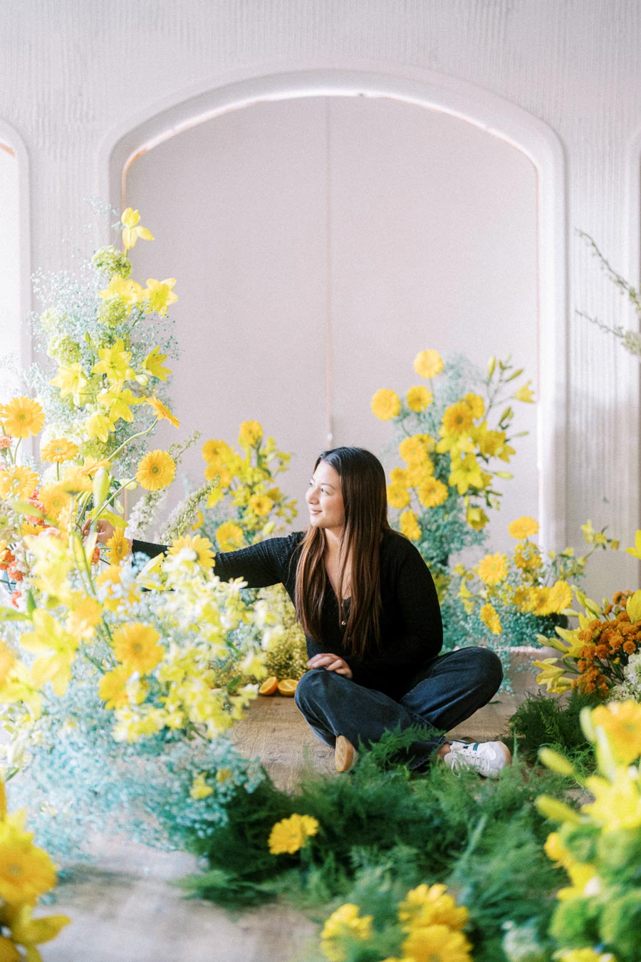 A woman arranging vibrant yellow and orange flowers in a bright room, surrounded by lush greenery, showcasing floral design and creativity.