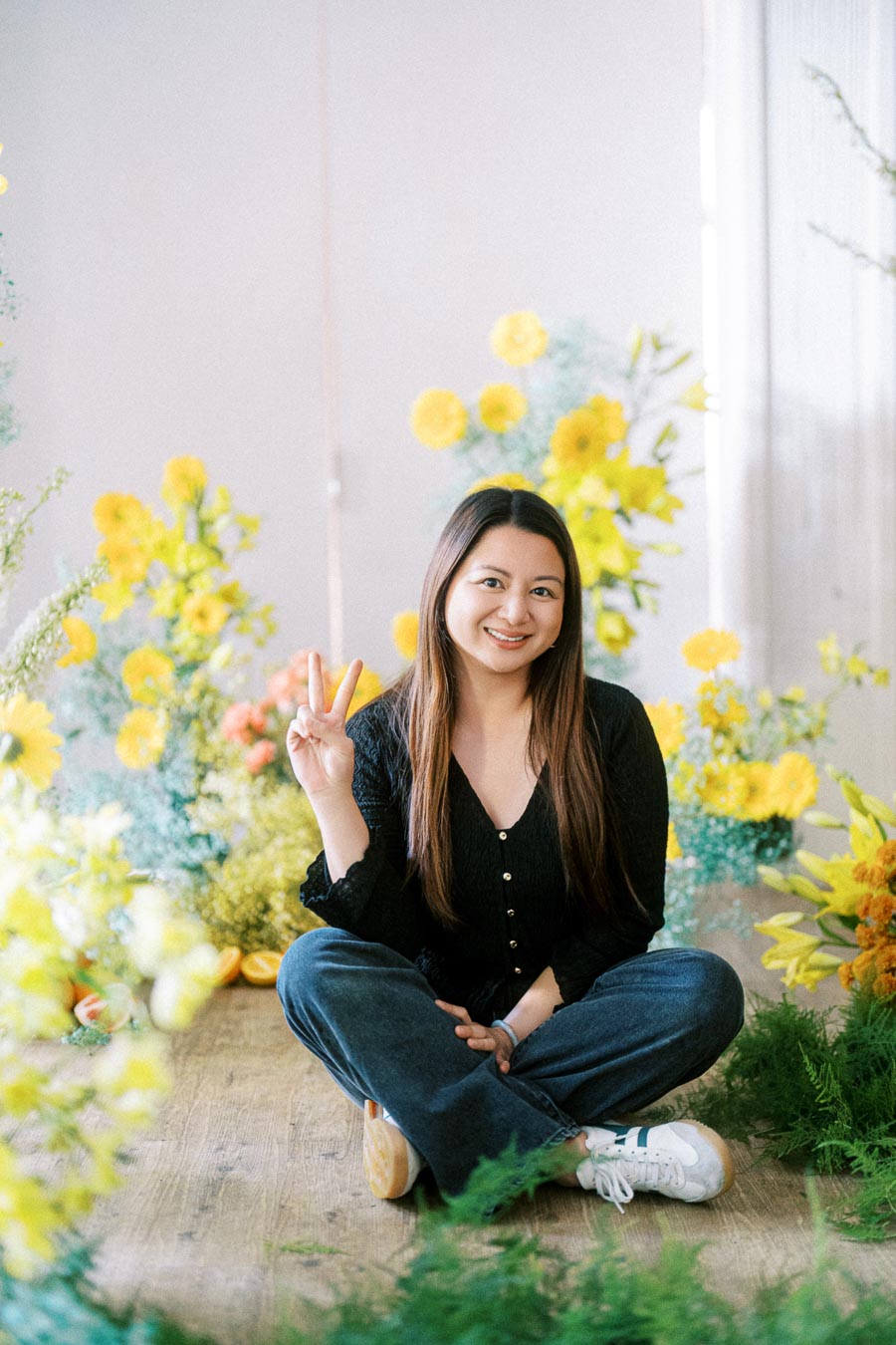 Smiling woman sitting on wooden floor surrounded by vibrant yellow flowers, making a peace sign with her fingers, wearing a black top and blue jeans.