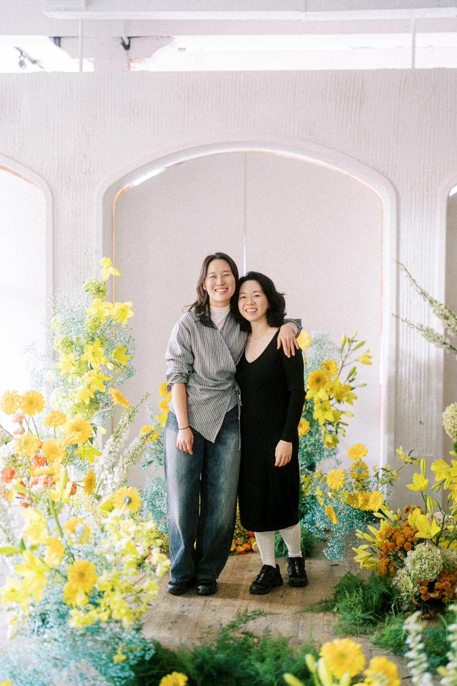 Two people smiling and embracing in front of a vibrant floral display with yellow and orange flowers, creating a cheerful and colorful atmosphere.