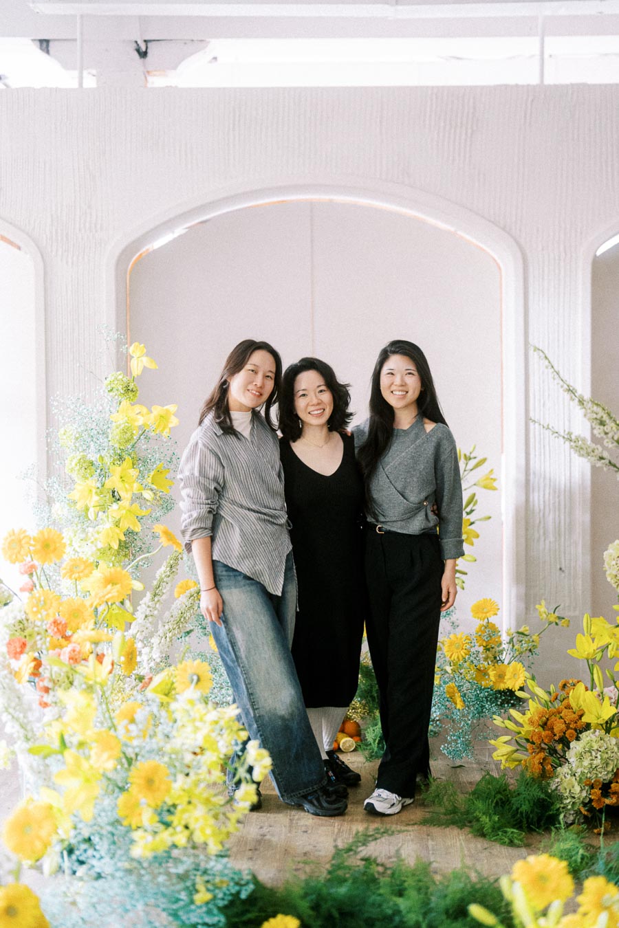 Three women smiling and posing together in front of a colorful floral backdrop, featuring yellow and orange blooms, in a bright indoor setting.