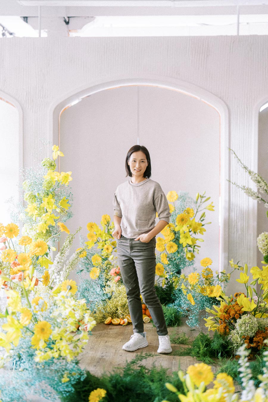 Person standing amidst vibrant yellow and green floral arrangement, wearing casual attire, with soft lighting enhancing the serene setting.