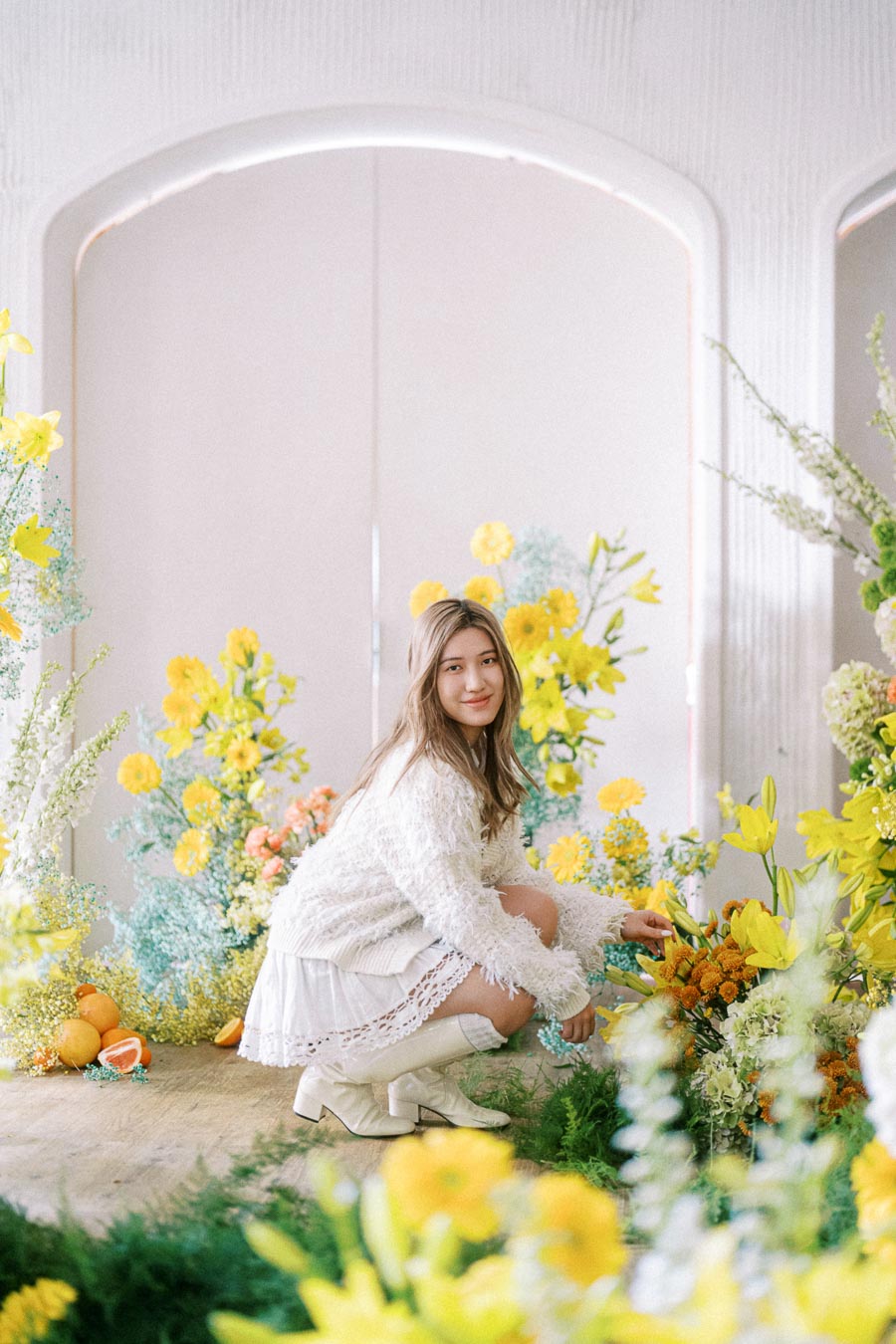 A woman in a white outfit crouches amid a vibrant arrangement of yellow and orange flowers, with a soft, natural light enhancing the serene and cheerful atmosphere.